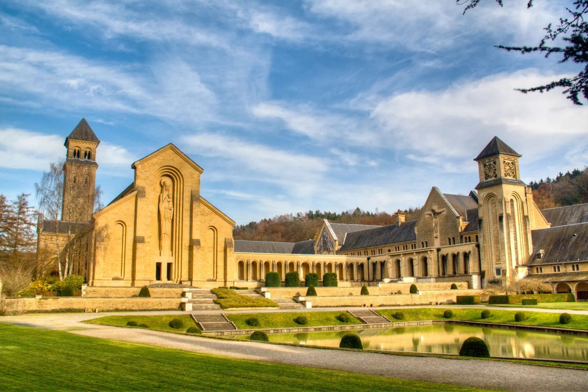 Blick auf die Zisterzienserabtei Orval mit imposanter Architektur aus hellem Stein, flankiert von zwei Türmen. Im Vordergrund befindet sich ein gepflegter Garten mit einem kleinen Teich. Der Himmel ist leicht bewölkt und die Umgebung ist von Bäumen umgeben.