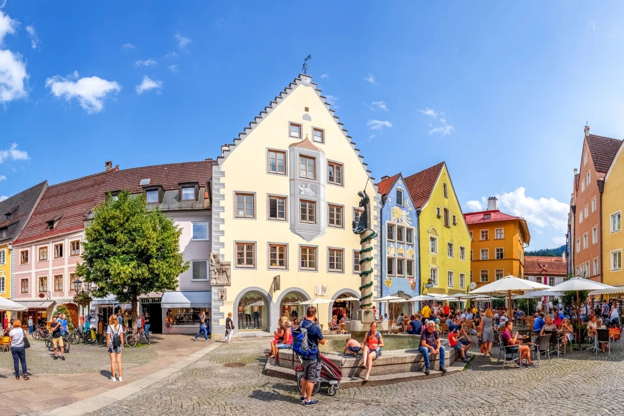 Das Bild zeigt den lebendigen Marktplatz in Füssen, umgeben von historischen Gebäuden mit bunten Fassaden, die typisch für die Altstadt der bayerischen Stadt sind. Auf dem Platz sind Menschen zu sehen, die in den Cafés entspannen, während Passanten durch die malerische Umgebung schlendern. Die einladende Atmosphäre wird durch das klare Wetter und die modernen Terrassenmöbel unter den Sonnenschirmen verstärkt. Im Hintergrund erheben sich die charakteristischen Dächer der traditionellen Häuser und eine markante Architektur ist ebenfalls gut erkennbar.