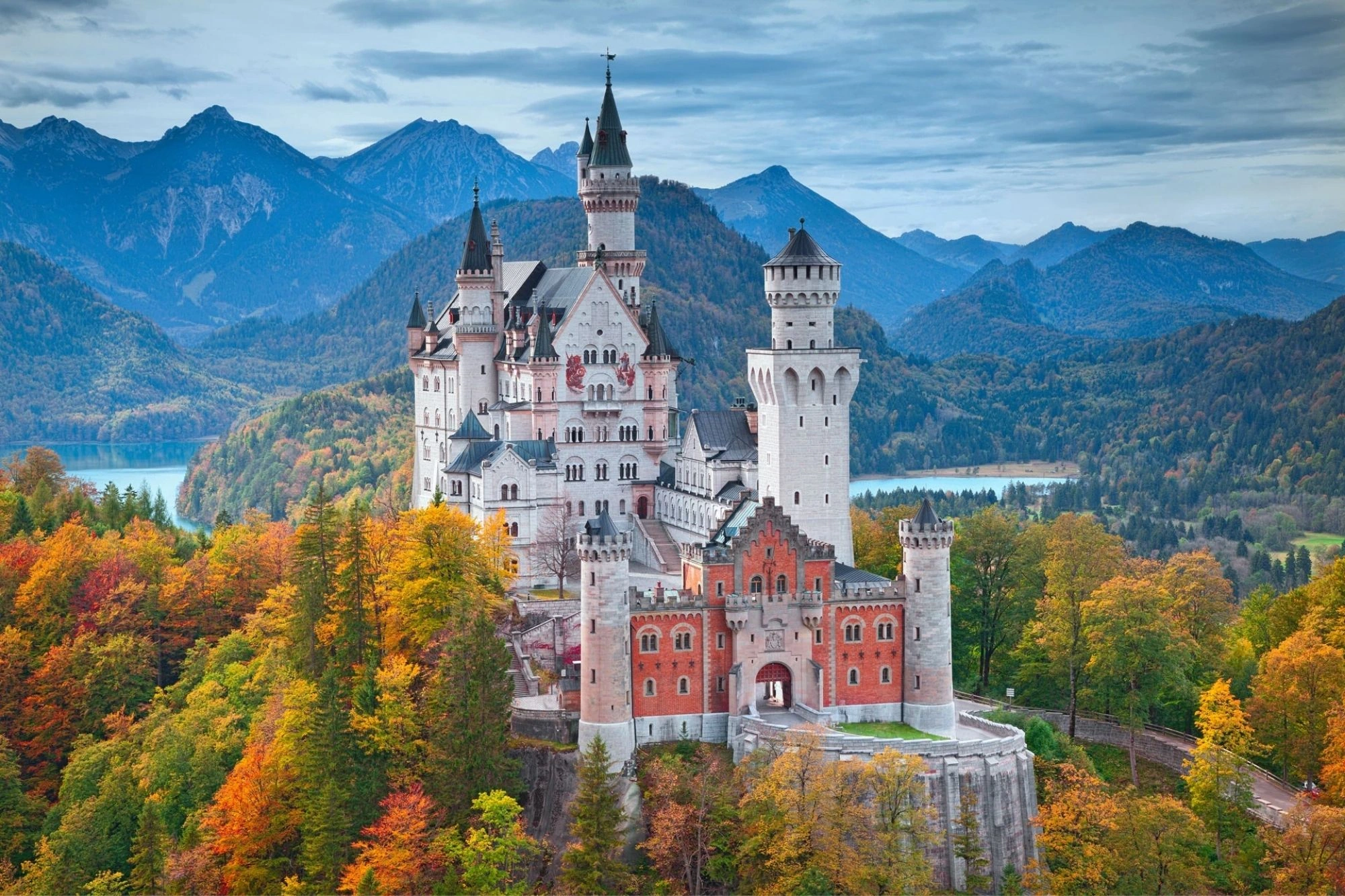 Das Bild zeigt Schloss Neuschwanstein, majestätisch auf einem Hügel gelegen, umgeben von herbstlich gefärbten Wäldern und einer beeindruckenden Alpenkulisse. Der malerische Bau mit seinen Türmen und Zinnen erhebt sich über die Landschaft, während im Hintergrund die blauen Berge und ein glitzernder See zu sehen sind. Diese Ansicht vermittelt eine romantische und traumhafte Atmosphäre, die das Schloss als eine der bekanntesten Sehenswürdigkeiten Deutschlands präsentiert.