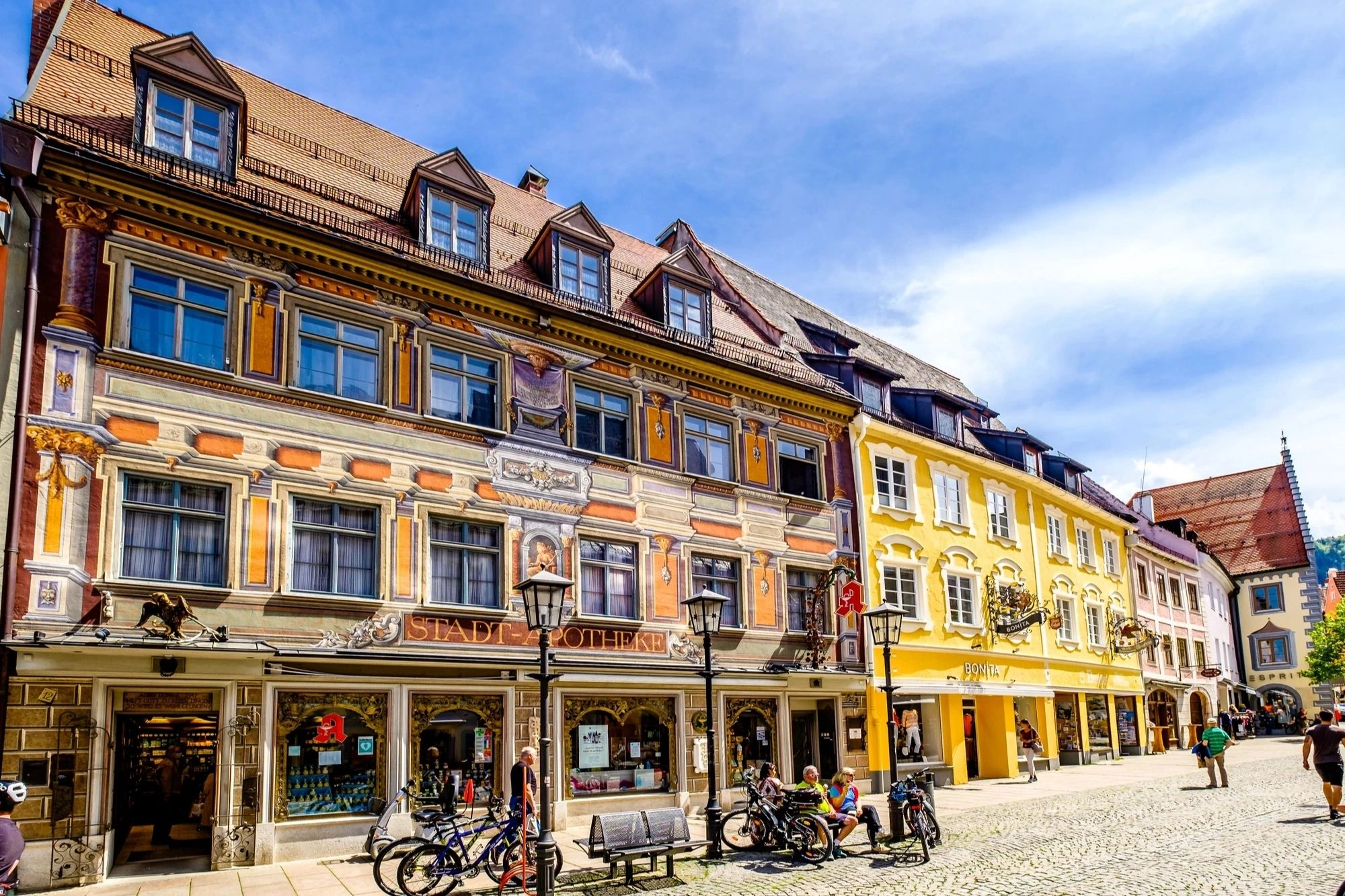 Das Bild zeigt eine malerische Straßenszene in Füssen, geprägt von historischen Gebäuden mit kunstvollen Fassaden, die in hellen Farbtönen erstrahlen. Die Stadt ist bekannt für ihre gut erhaltene Altstadt, die von charmanten Geschäften und Cafés gesäumt wird. Im Vordergrund sind mehrere Fahrräder abgestellt, während Passanten die Straße entlang flanieren. Die Architektur spiegelt die Tradition und den Charakter dieser bayerischen Stadt wider, während das schöne Wetter und der klare Himmel eine einladende Atmosphäre schaffen.