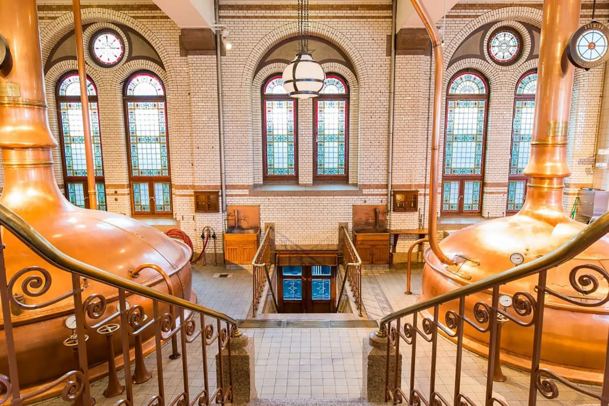 Das Bild zeigt eine Treppe im Heineken Museum in Amsterdam, die hinunter in den historischen Braukeller führt. Die Umgebung ist geprägt von großen, glänzenden Kupferkesseln, die traditionell für den Brauprozess verwendet wurden. Die hohen Fenster mit buntem Glas und die dekorativen Geländer verleihen dem Raum ein klassisches und stilvolles Ambiente, das die Geschichte und Tradition der Heineken-Brauerei widerspiegelt.