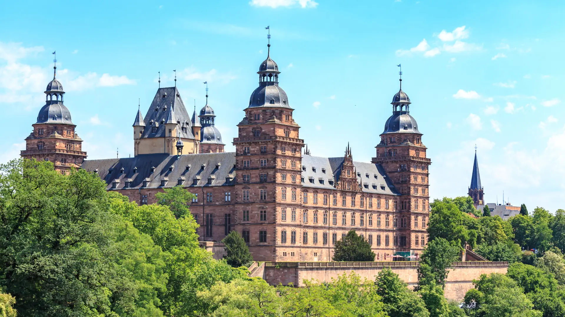 Das Bild zeigt das Schloss Johannisburg von außen. Es hat vier Türme aus rotem Sandstein und ist von grünen Bäumen umgeben, unter einem blauen Himmel.