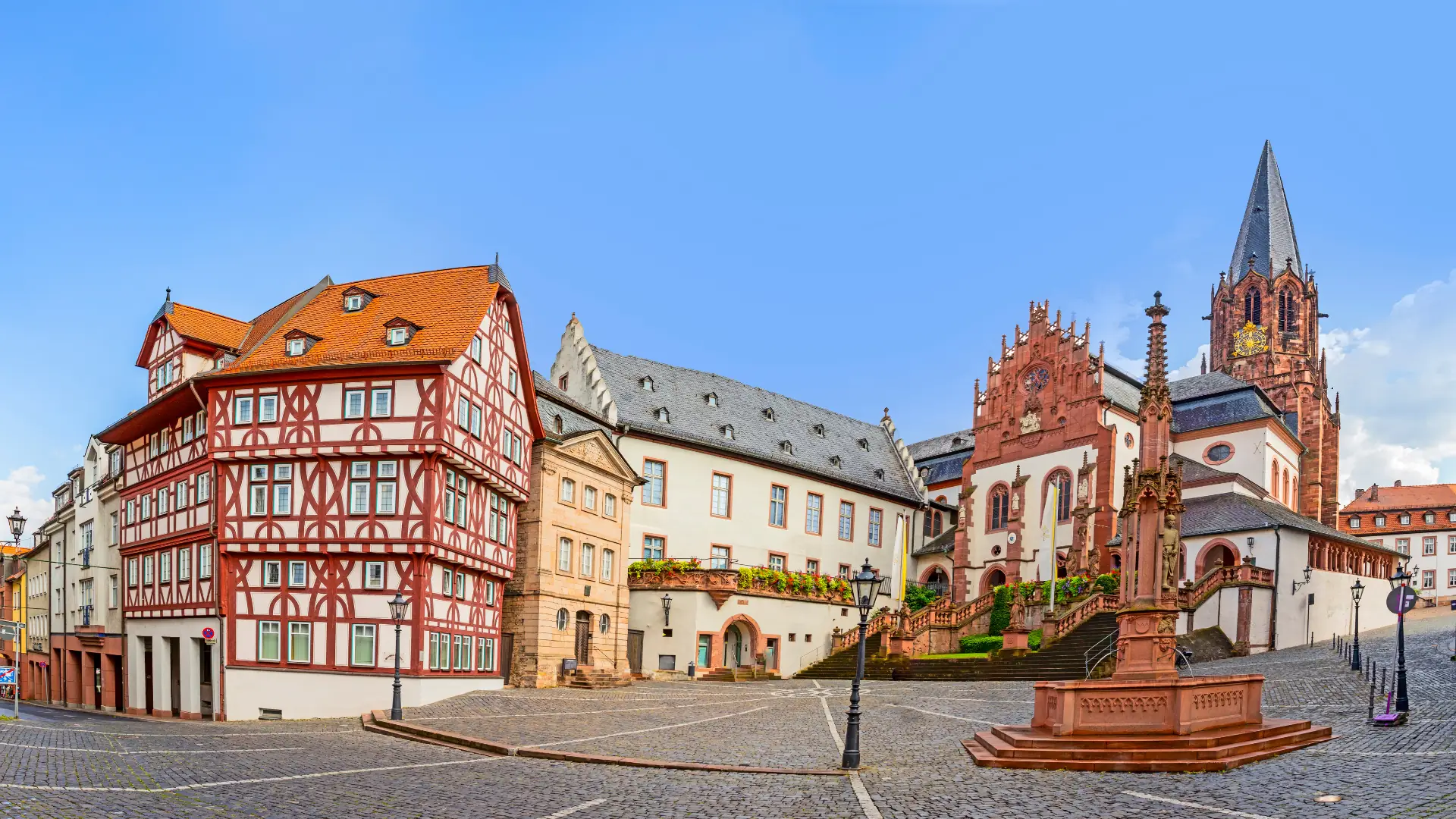 Das Bild zeigt den Marktplatz in Aschaffenburg mit historischen Fachwerkhäusern und gotischer Kirche mit hohem Turm auf einem gepflasterten Platz.