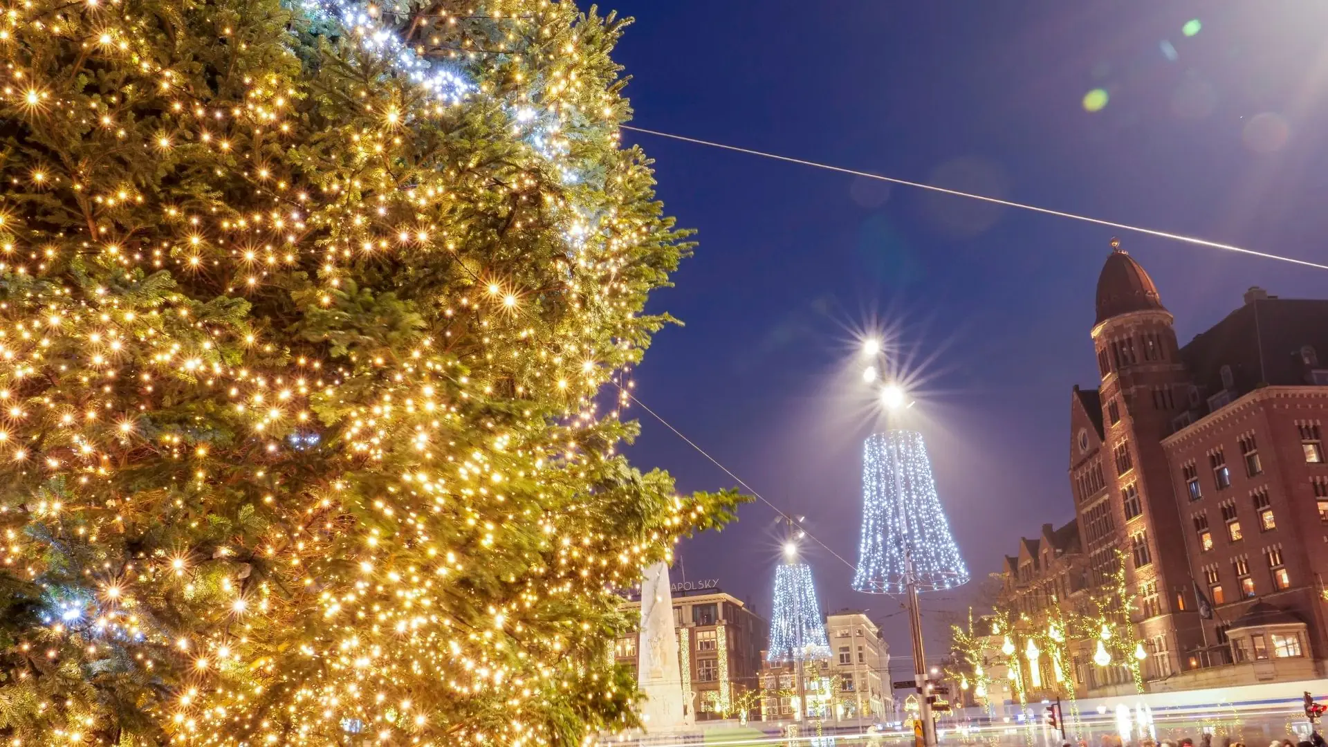 Beleuchteter Weihnachtsbaum links, mit Lichtern geschmückt. Im Hintergrund festliche Lichter in Form von Glocken und historische Gebäude in Amsterdam.