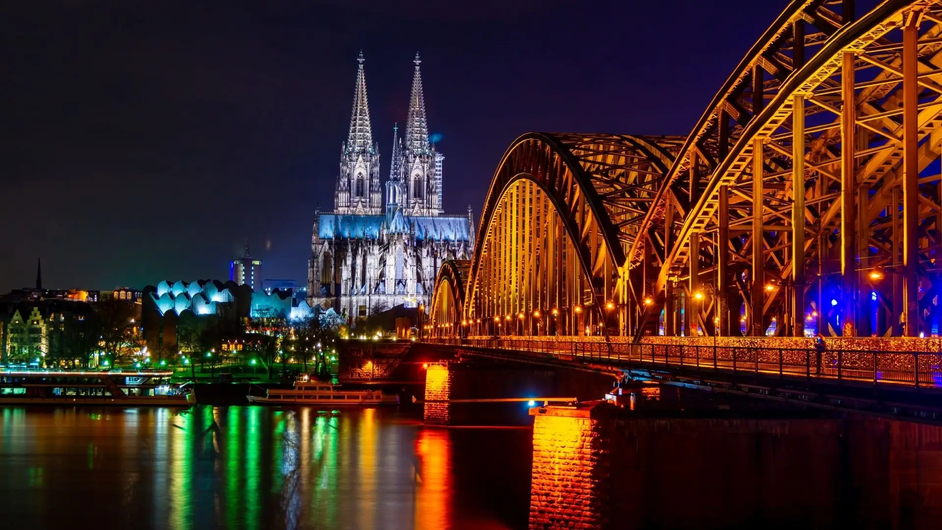 Kölner Dom mit zwei Türmen im Hintergrund, beleuchtet bei Nacht. Im Vordergrund die Hohenzollernbrücke mit orangefarbenen Lichtern und reflektierenden Wasserflächen.