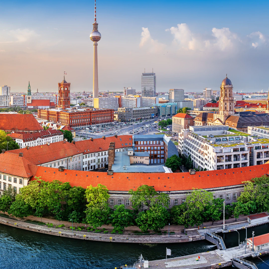Das Bild zeigt eine Panorama-Ansicht von Berlin bei Sonnenuntergang. Im Vordergrund fließt die Spree mit mehreren Brücken, während historische Gebäude mit roten Dächern das Stadtbild prägen. Der Berliner Fernsehturm ragt markant in die Höhe, daneben sind das Rote Rathaus und der Berliner Dom zu sehen. Die Skyline kombiniert moderne Hochhäuser mit klassischer Architektur. Der Himmel ist in warmen Farben gefärbt und verleiht der Szene eine atmosphärische Tiefe.