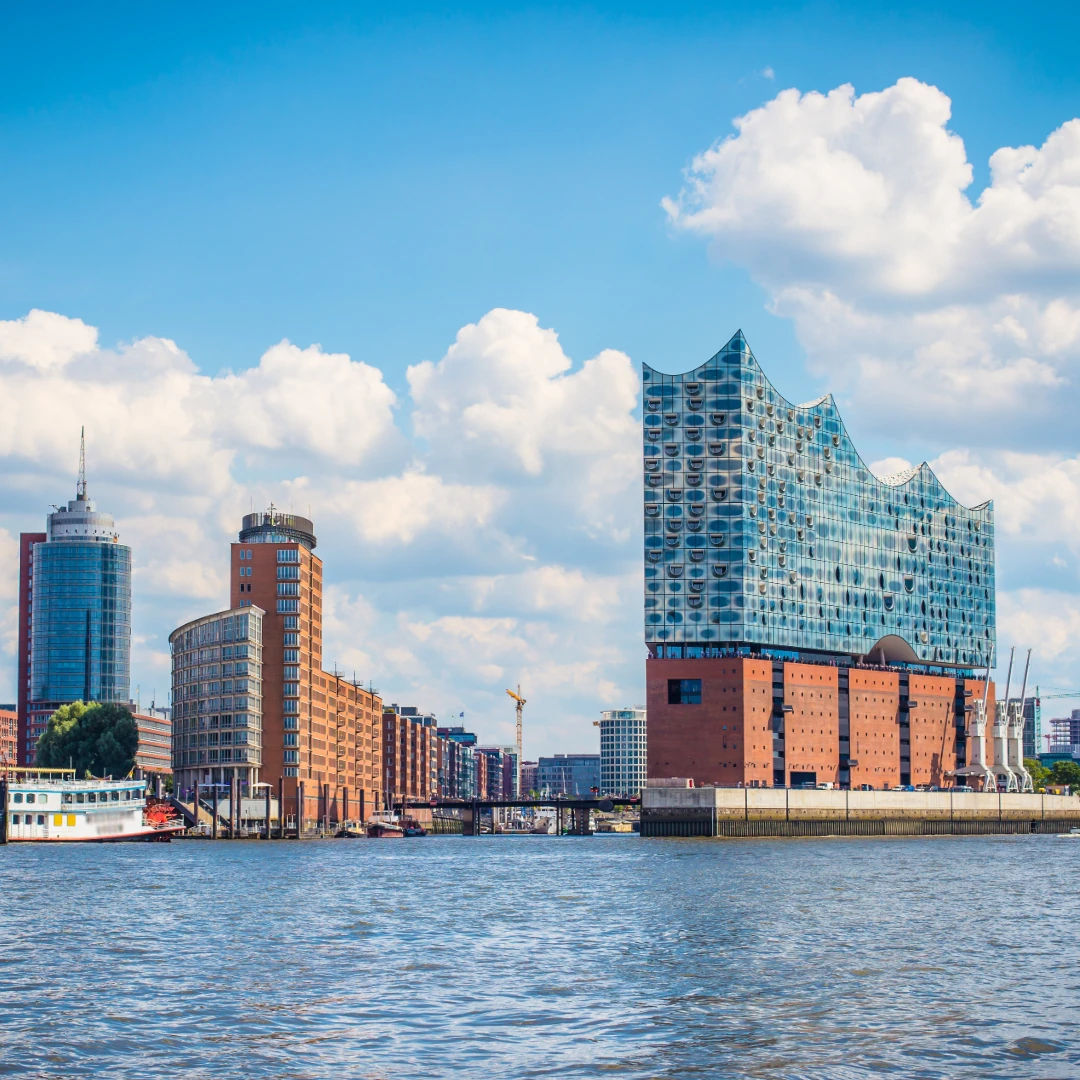 Das Bild zeigt die Hamburger Elbphilharmonie, eines der markantesten Wahrzeichen der Stadt, mit ihrer wellenförmigen Glasfassade über dem historischen Kaispeicher. Im Vordergrund fließt die Elbe, während ein klassisches Schiff am Ufer liegt. Die Skyline kombiniert moderne Hochhäuser mit traditioneller Hafenarchitektur unter einem Himmel mit weißen Wolken.