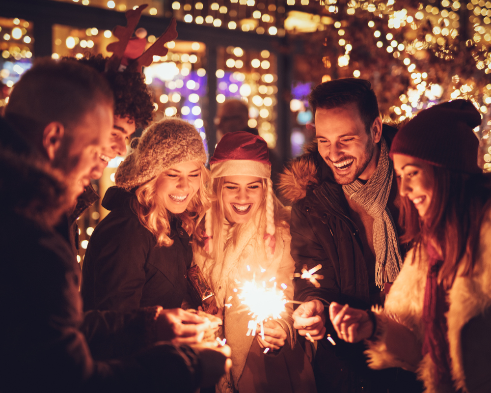 Das Bild zeigt eine fröhliche Gruppe von Menschen, die gemeinsam bei festlichen Lichtern und funkelnden Feuerwerkslichtern lachen und eine wunderbare Zeit genießen. Alle tragen winterliche Kleidung und festliche Accessoires wie Weihnachtsmützen und -ohren. Die Szene vermittelt eine warme und einladende Atmosphäre, die mit dem Glanz der Lichter und der ausgelassenen Stimmung der Teilnehmer zu einer fröhlichen Feier beiträgt.