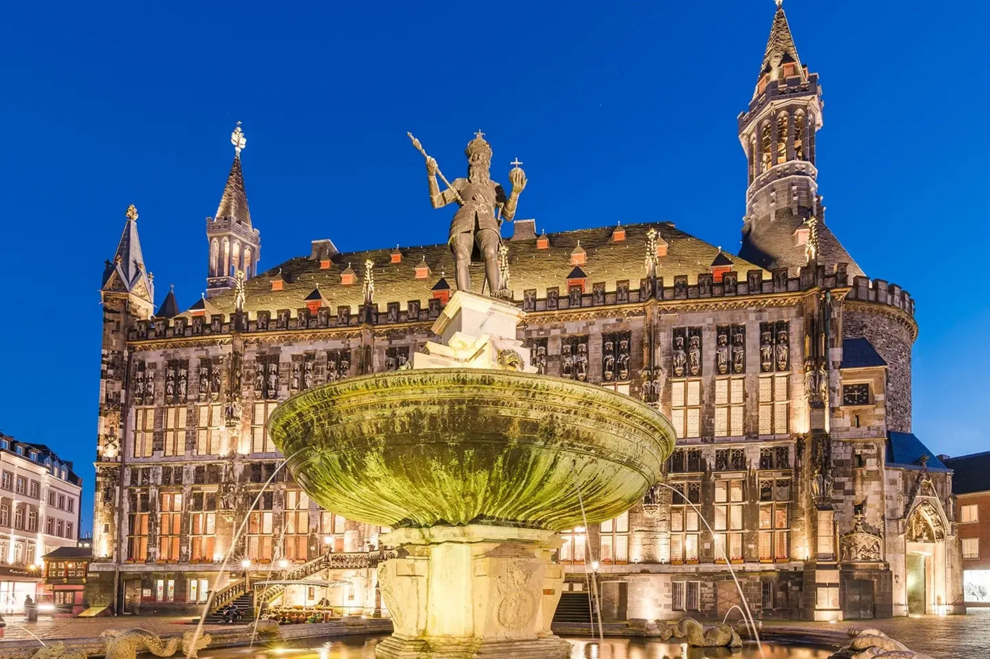 Neptunbrunnen mit beleuchtetem historischen Rathaus im Hintergrund bei Abenddämmerung in Aachen