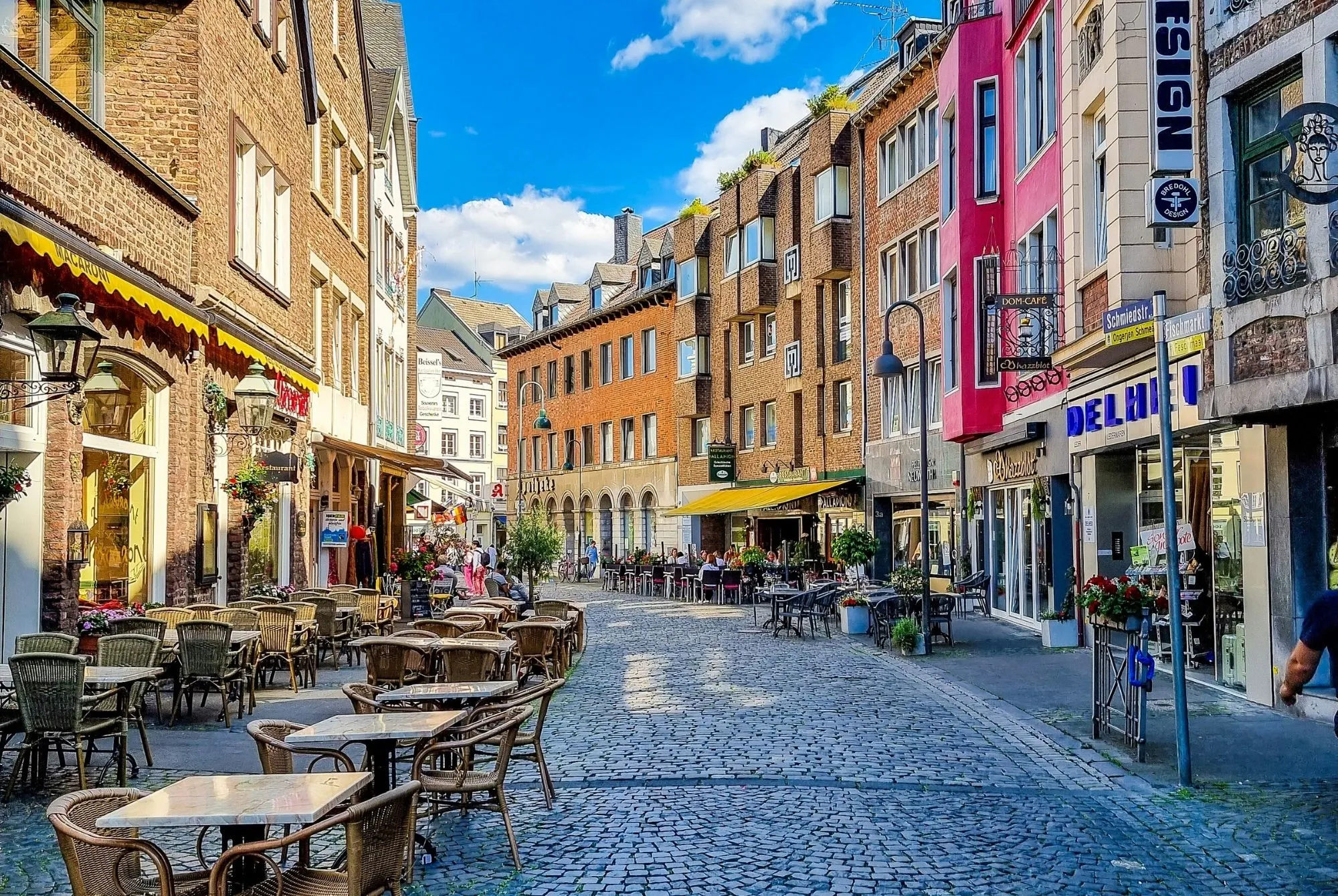Pflasterstraße mit Cafés und Restaurants, Tische und Stühle im Freien, mehrstöckige Gebäude mit Geschäften und Wohnungen, blauer Himmel