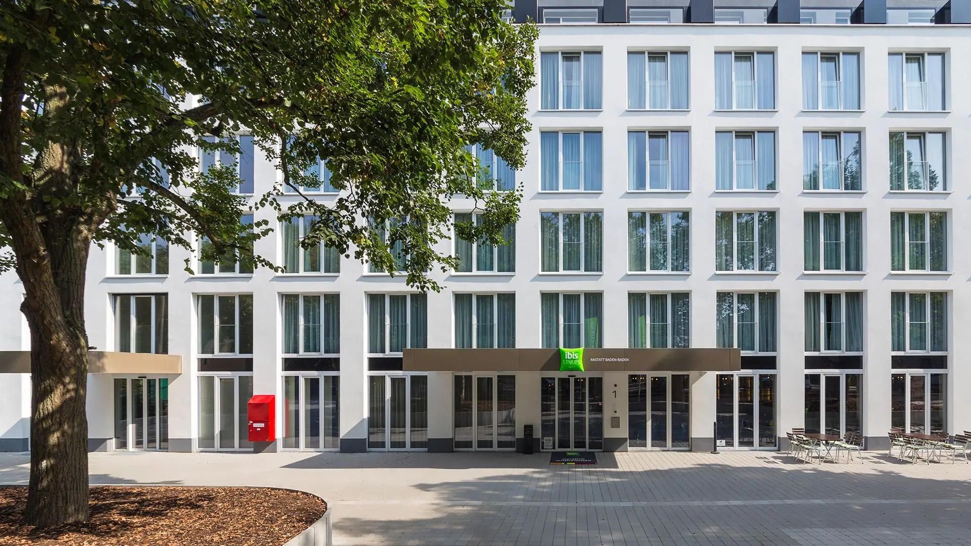Eingang des ibis Hotels mit moderner Fassade, großen Fenstern und Sitzbereich im Freien
