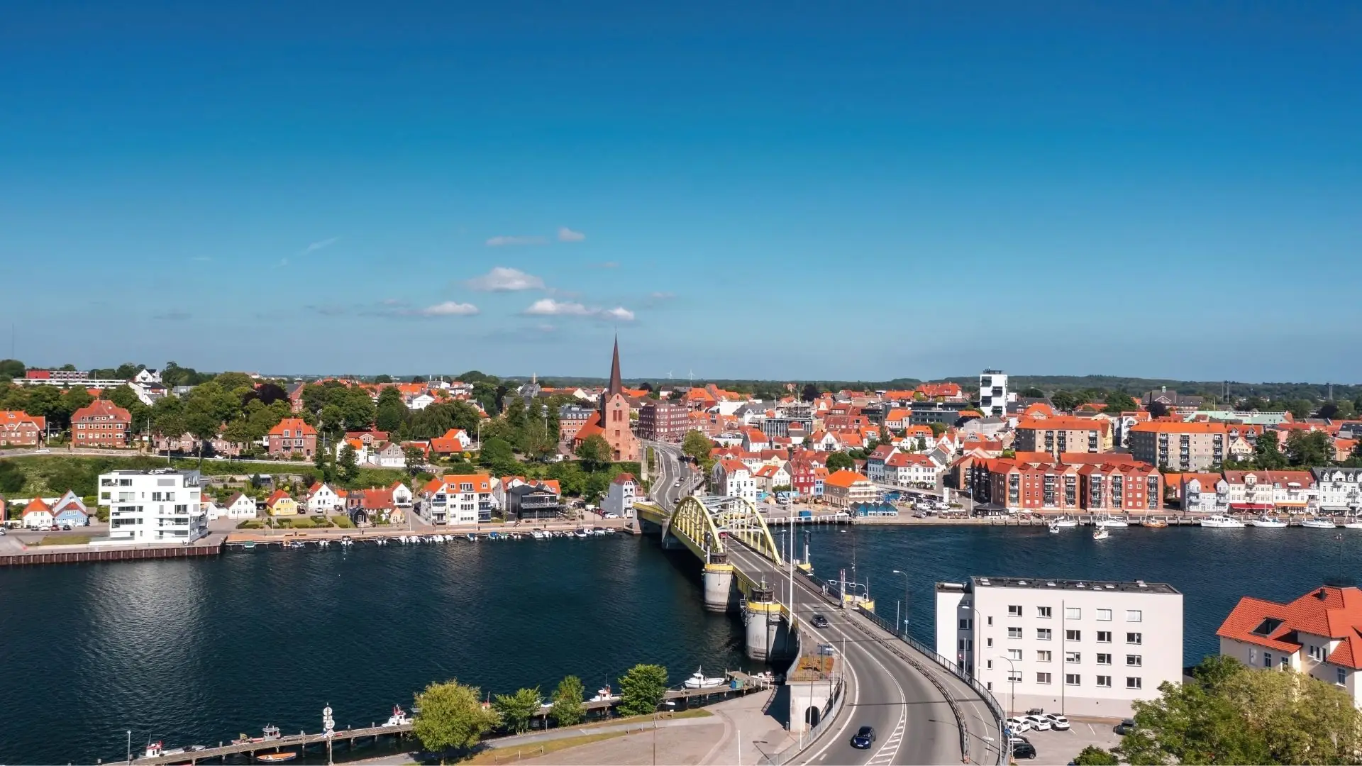 Luftaufnahme von Sonderburg mit Brücke über den Fluss, roten Ziegeldächern, Kirche mit hohem Turm und blauem Himmel.