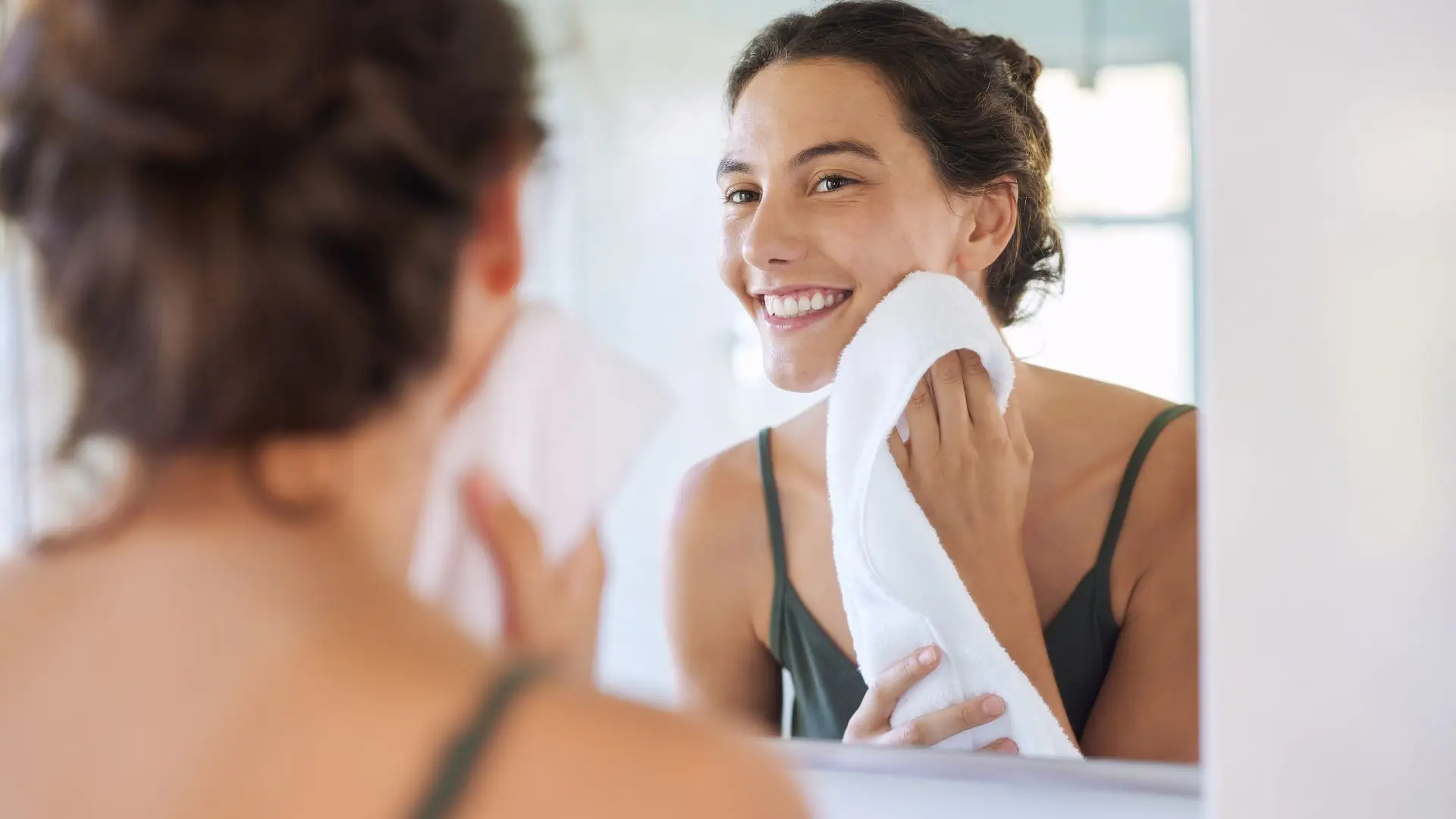 Frau mit hochgestecktem Haar in grünem Trägeroberteil trocknet ihr Gesicht mit weißem Handtuch vor einem Spiegel