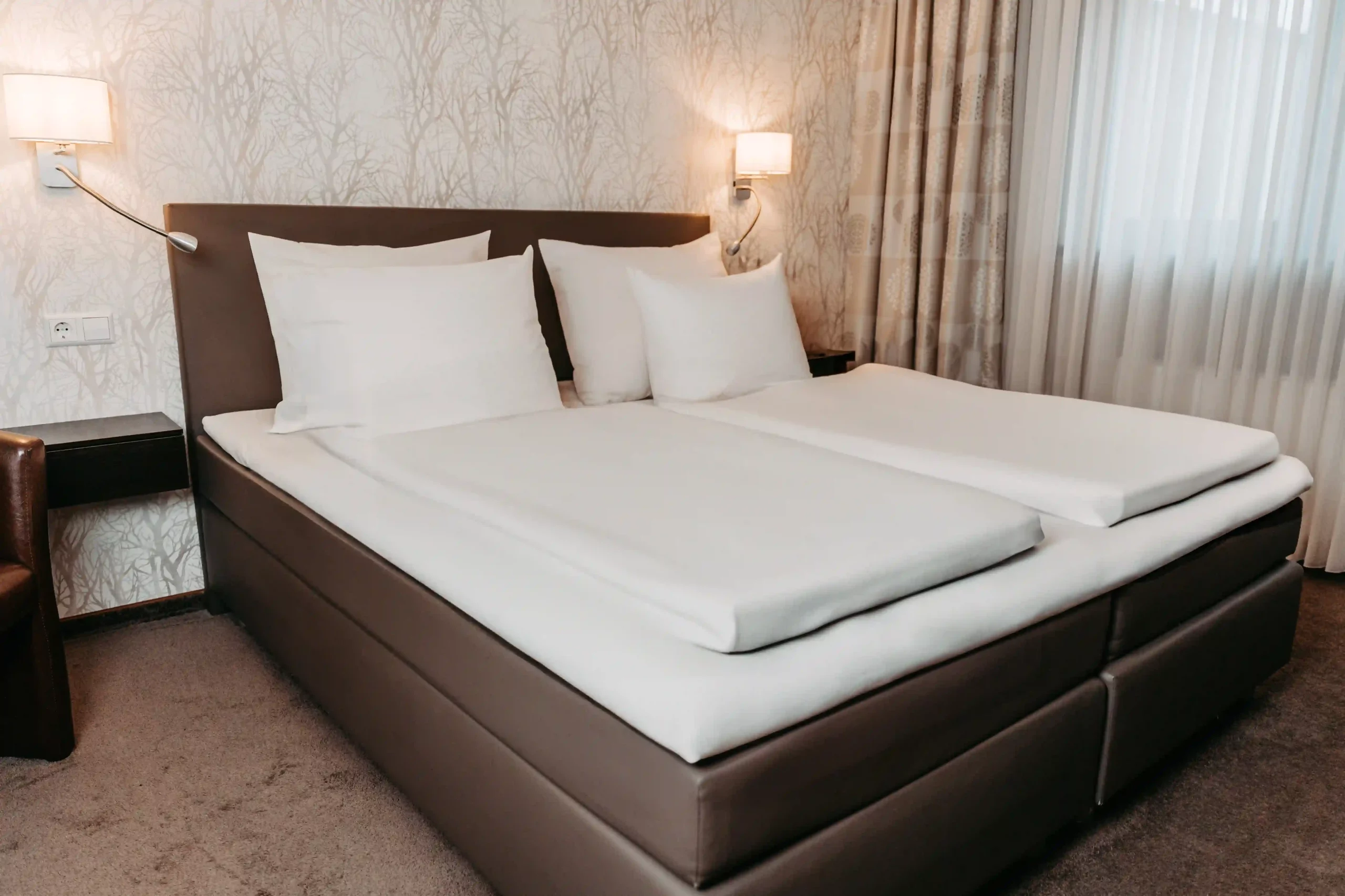 Doppelbett mit braunem Lederbettgestell, weißer Bettwäsche und mehreren Kissen in einem Hotelzimmer mit gemusterter Tapete, Vorhängen und zwei Wandlampen