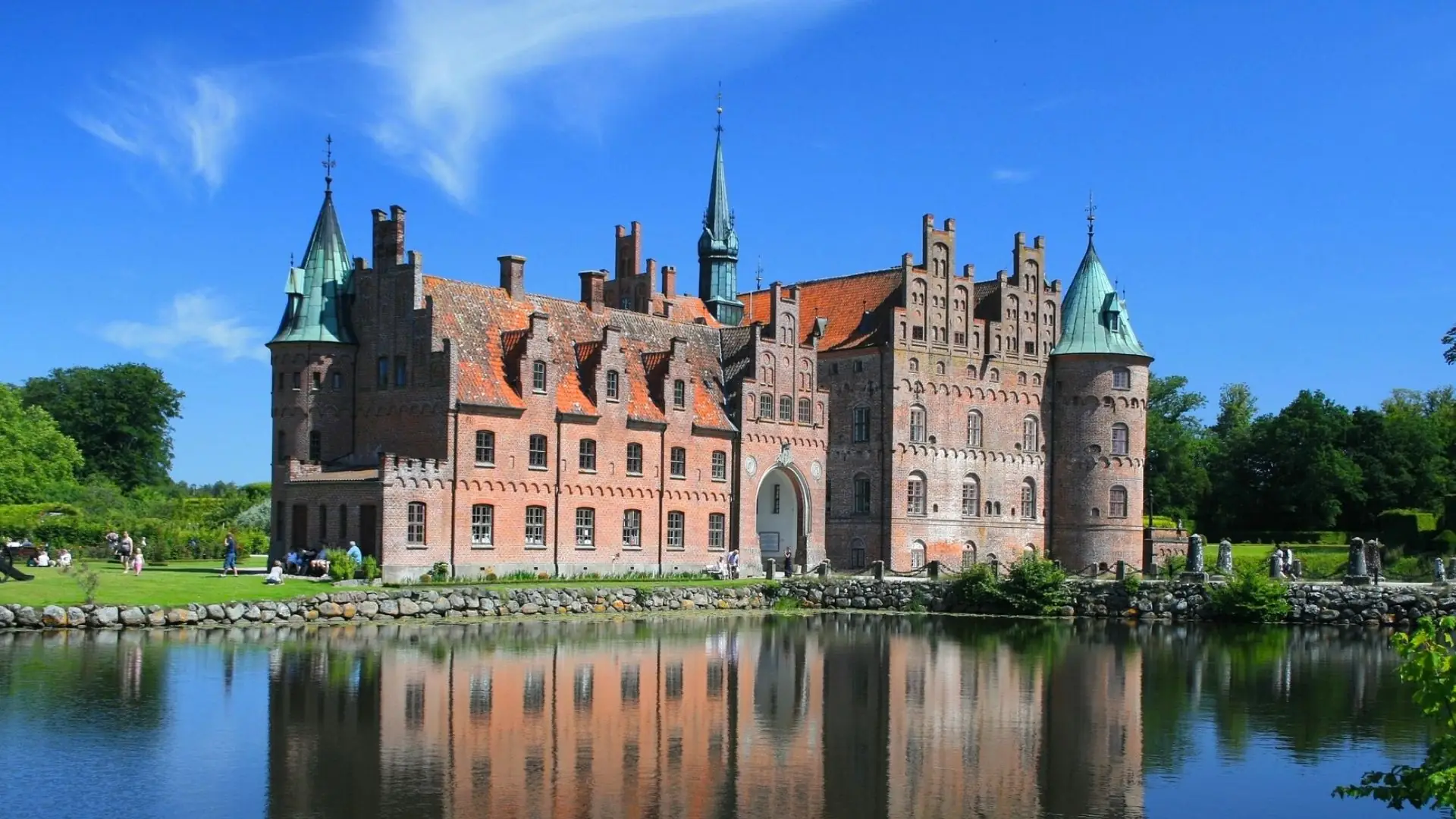 Egeskov Schloss in Odense mit roten Ziegelmauern, Türmen mit grünen Dächern und Spiegelung im Wasser unter blauem Himmel.