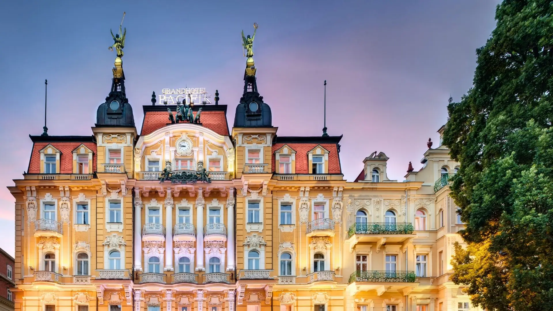 Außenansicht des historischen Grandhotel Pacifik mit zwei vergoldeten Figuren auf den Türmen und beleuchteter Fassade bei Abenddämmerung.