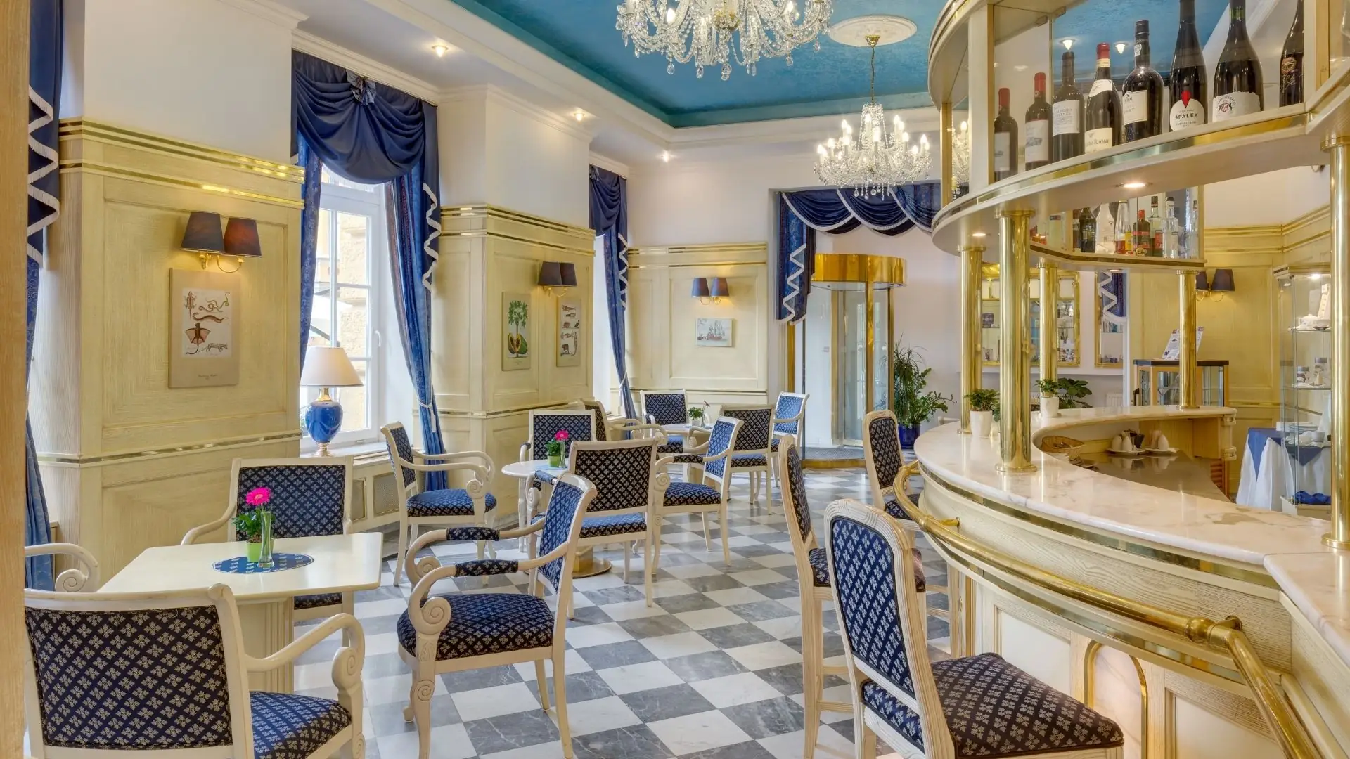 Elegante Hotellobby mit blau gepolsterten Stühlen, weißen Tischen, Kronleuchtern und einer Bar mit Weinflaschen.