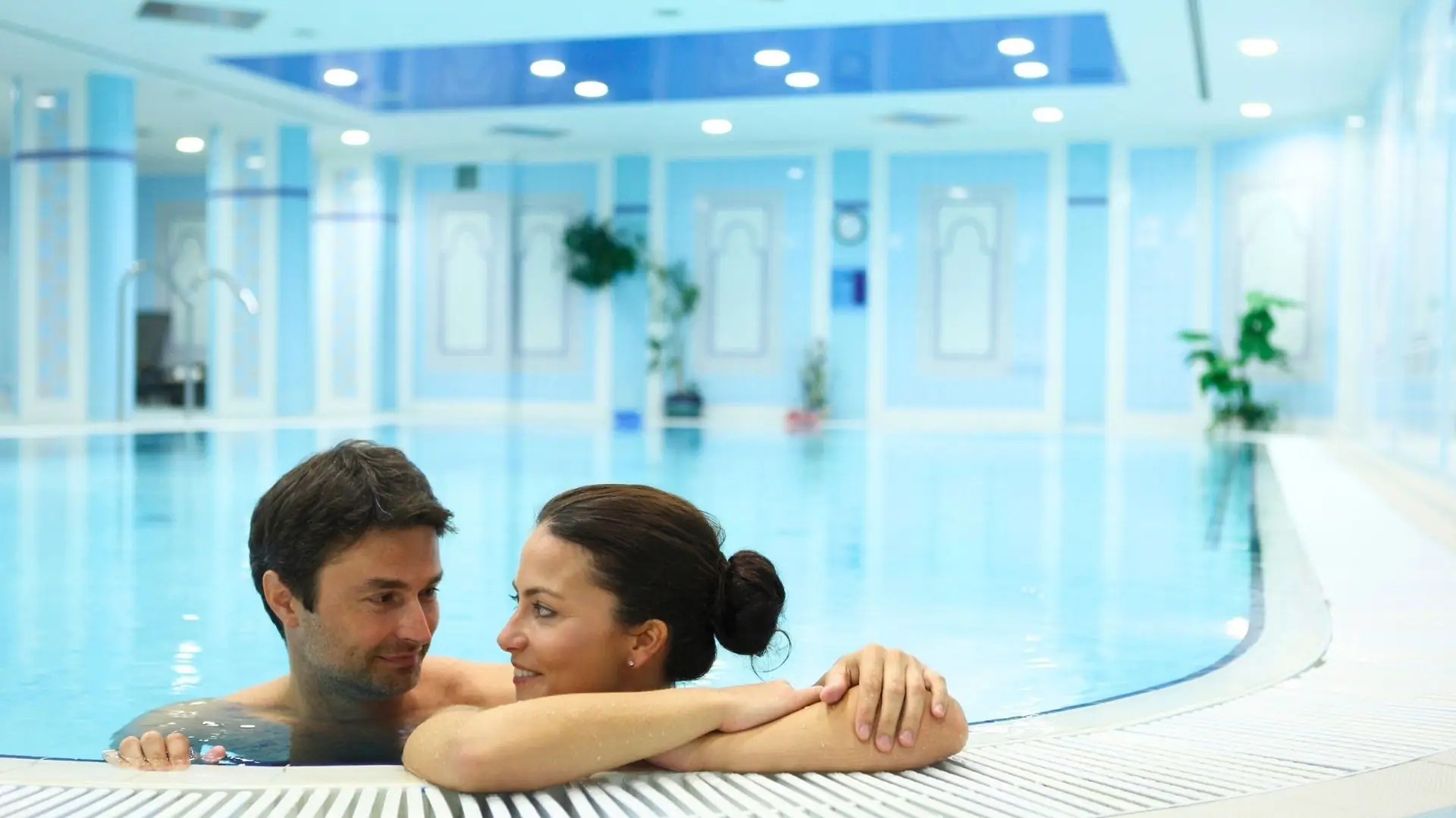 Paar entspannt am Beckenrand eines hellen Innenpools im Hotel Pacifik, umgeben von blauen Wänden und Pflanzen.