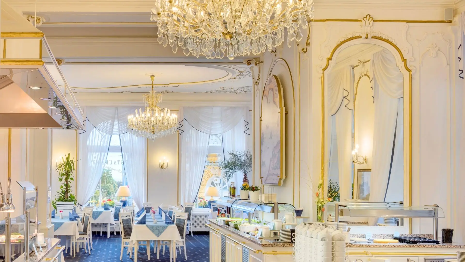 Elegantes Restaurant mit Kronleuchtern, großen Fenstern, weißen Tischdecken und goldverzierten Spiegeln an den Wänden.
