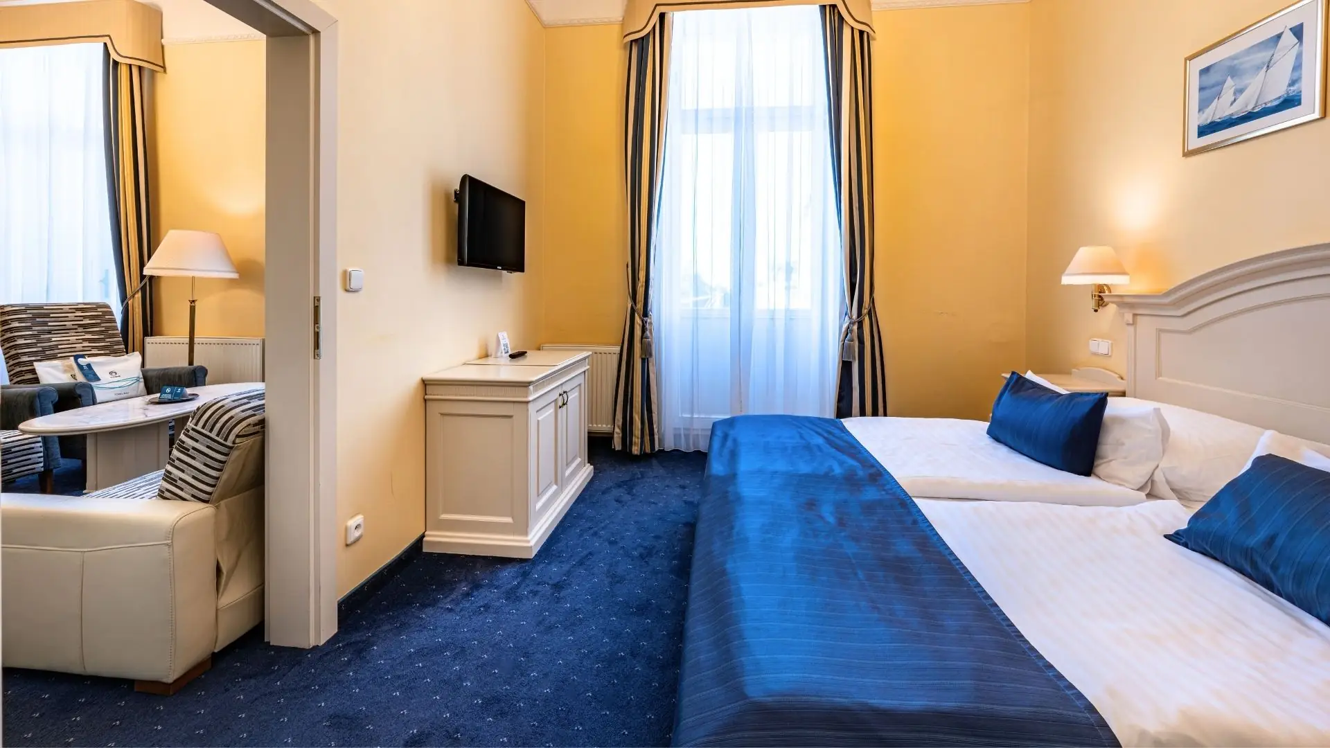 Hotel Pacifik Superior Doppelzimmer, mit Doppelbett, blauer Bettwäsche und Teppich, Sofa mit Kissen, Wandspiegel, Flachbildfernseher und Fenster mit Vorhängen.