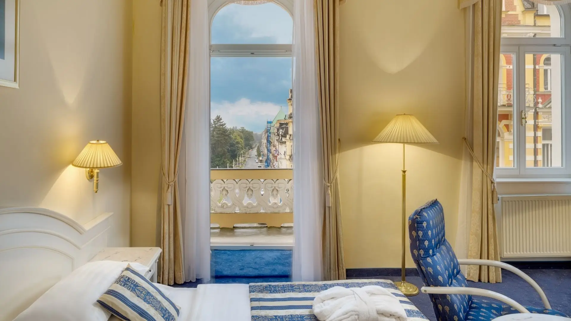 Hotelzimmer mit Doppelbett, blau-weiß gestreiften Kissen und Decke, Blick durch offene Balkontür auf Straße und Gebäude, zwei Lampen an Wand und Stehlampe, blaue Teppichboden.