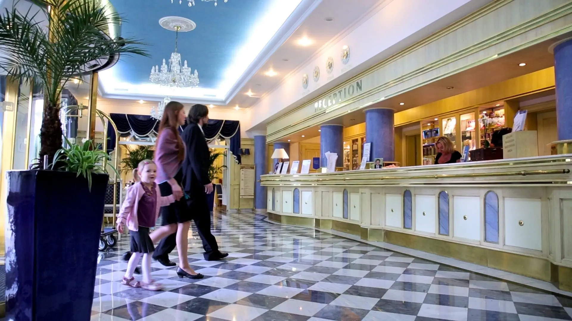 Hotelrezeption mit Marmorfußboden, Kronleuchter und mehreren Personen, darunter zwei Erwachsene und zwei Kinder, die vorbeigehen.
