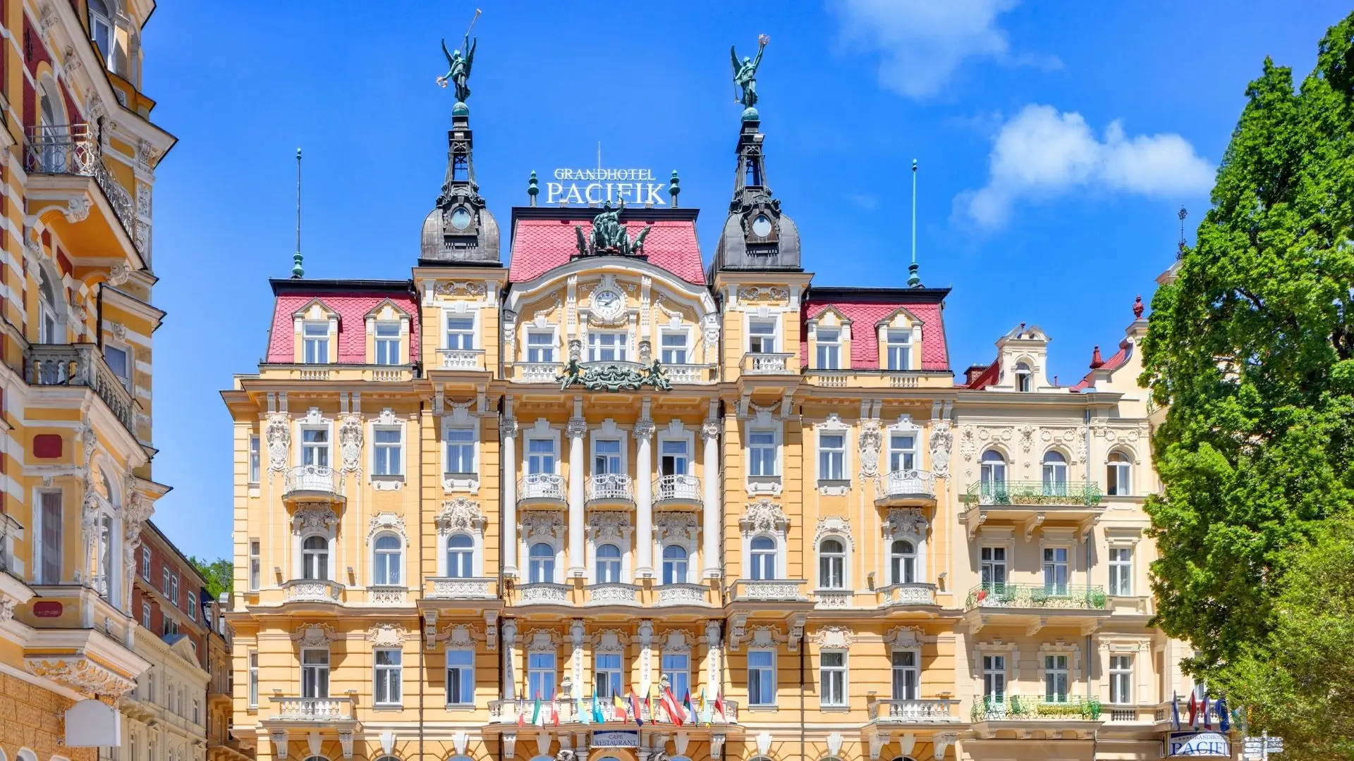 Fassade des Grandhotel Pacifik mit reich verzierten Balkonen, zwei Statuen auf dem Dach und mehreren Flaggen vor dem Eingang.