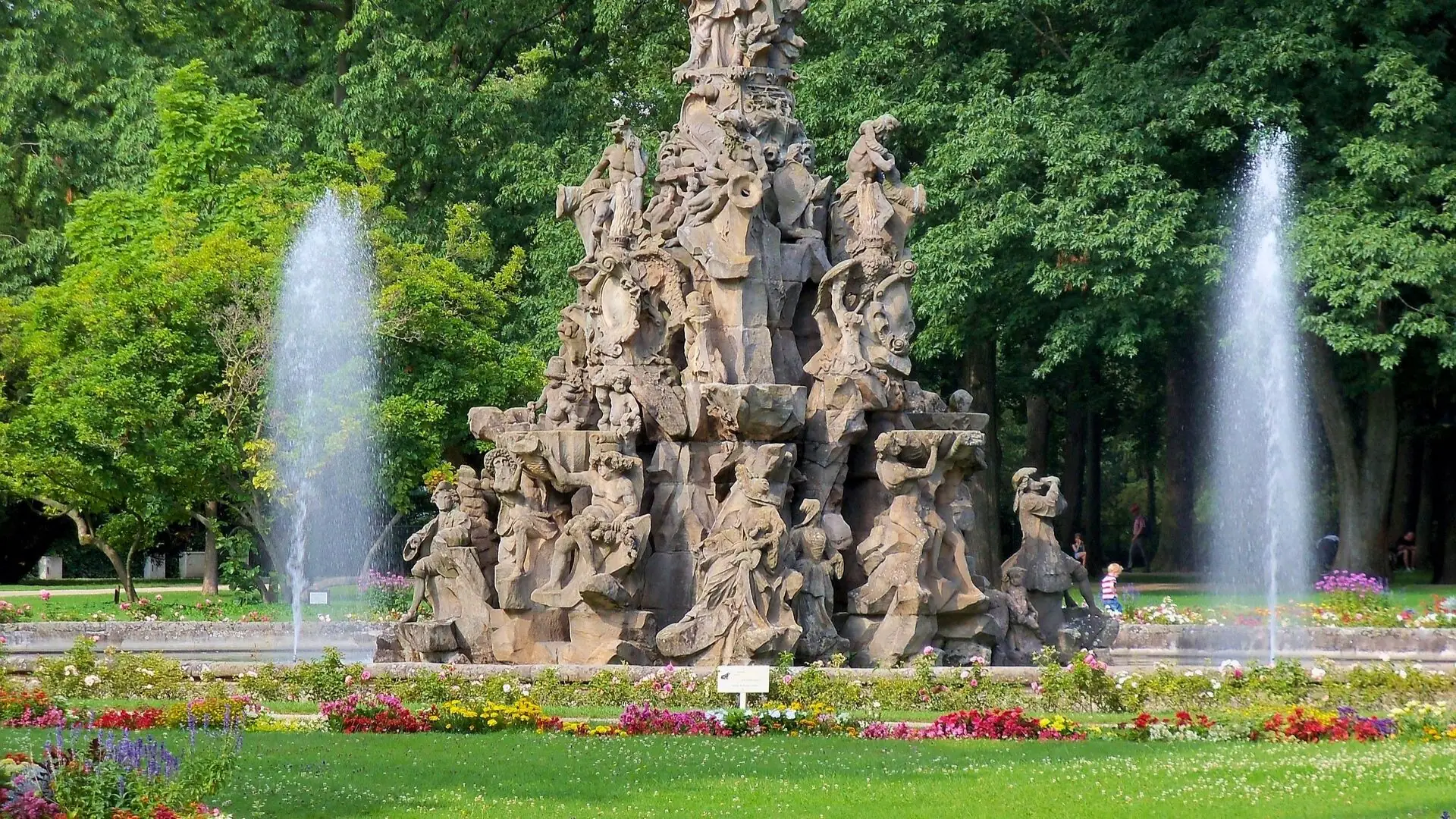 Brunnen mit mehreren Skulpturen von Menschen und Tieren, umgeben von Wasserfontänen und bunten Blumenbeeten in einem Park.