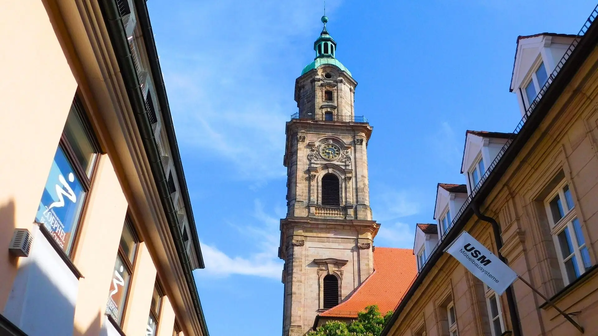 Blick auf den Turm der Neustädterkirche in Erlangen, umgeben von Gebäuden mit Fenstern und einem USM-Schild.