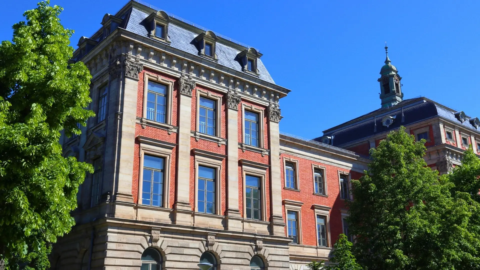 Historisches Gebäude der Universität Erlangen mit roter und grauer Fassade, verzierten Fenstern und einem Turm im Hintergrund.