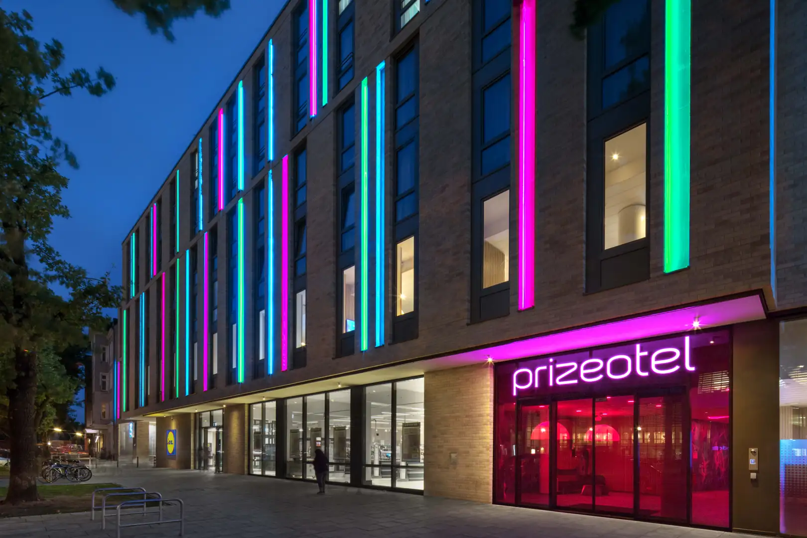 Helle und moderne Fassade des prizeotel bei Nacht mit bunten Lichtern.