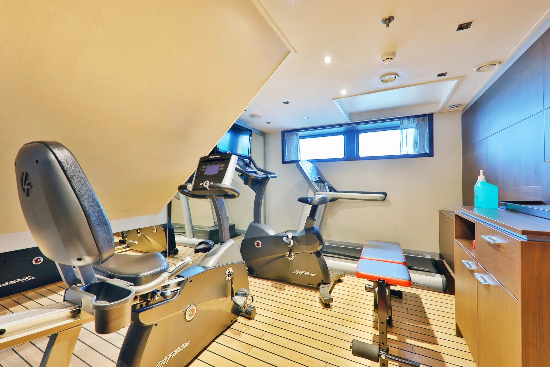 Kleiner Fitnessraum mit zwei Ergometern, einer Hantelbank und einem Fenster unter der Decke