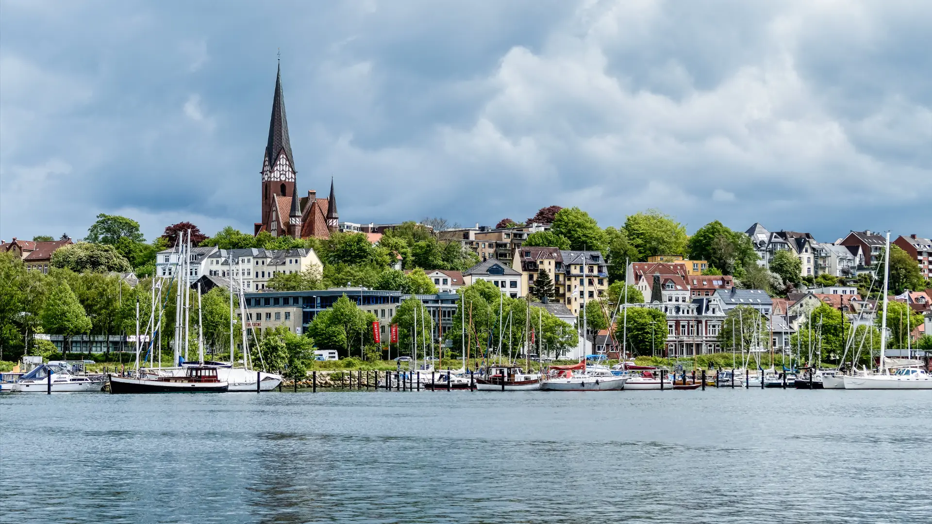 Blick auf Flensburg vom Wasser aus mit Segelbooten im Hafen und der St. Jürgen-Kirche im Hintergrund.
