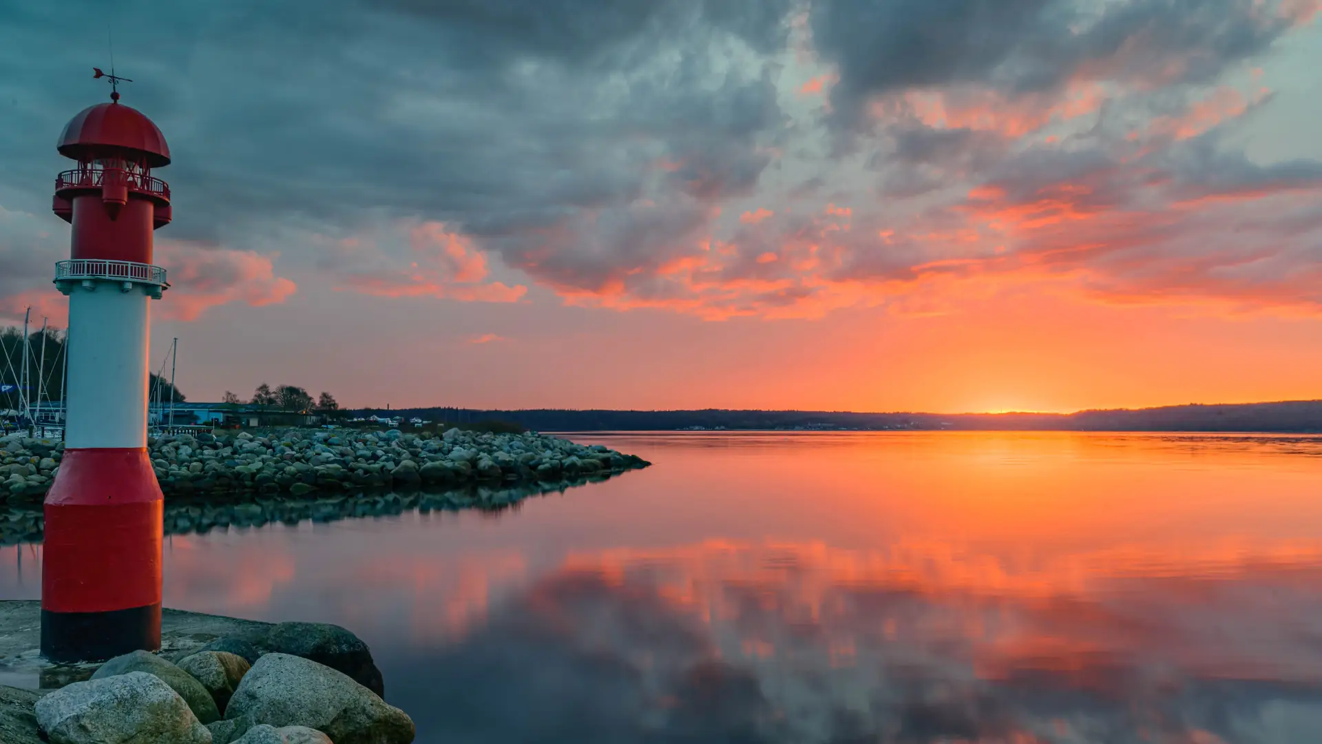 Das Bild zeigt einen Leuchtturm mit rotem und weißem Streifen am Ufer, ruhiges Wasser und bunter Sonnenuntergangshimmel.