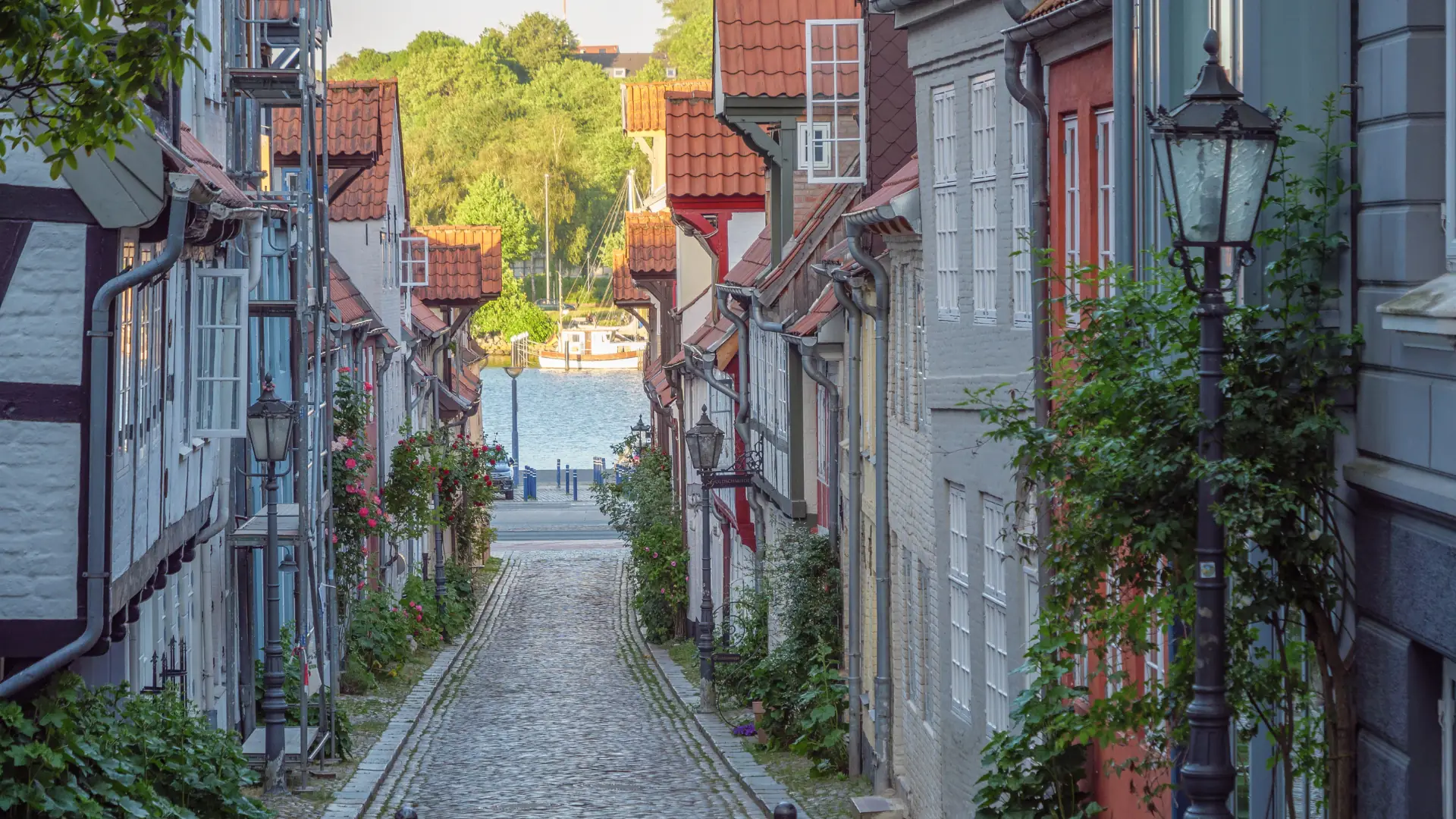 Das Bild zeigt eine schmale Kopfsteinpflasterstraße mit alten Häusern und Laternen, am Ende Blick auf einen Fluss mit Booten.