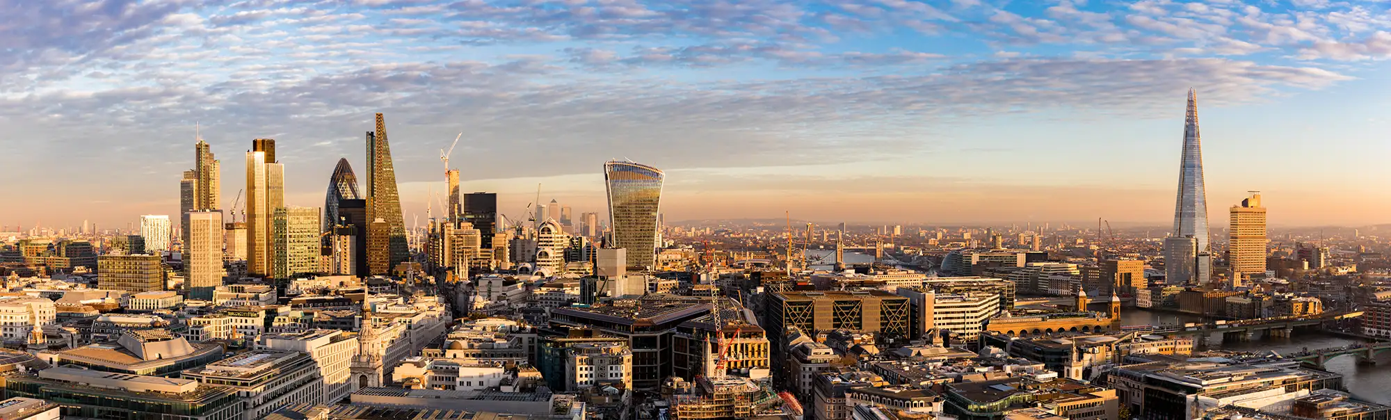 Die Skyline von London ist eine atemberaubende Mischung aus moderner Architektur und historischen Gebäuden. In dieser Panoramaaufnahme sind einige der bekanntesten Wolkenkratzer und Wahrzeichen der Stadt zu sehen. Der Shard, mit seiner markanten spitzen Form, sticht als eines der höchsten Gebäude der Stadt hervor. Daneben sind mehrere andere moderne Bürogebäude sichtbar, die zusammen ein faszinierendes Stadtbild schaffen. Das Bild zeigt auch die Themse, die sich durch die Stadt schlängelt, und die warmen Farbtöne des Sonnenuntergangs, die die Szene in ein sanftes Licht tauchen. Die Kombination aus Alt und Neu in dieser urbanen Landschaft verkörpert das dynamische und vielfältige Wesen Londons.