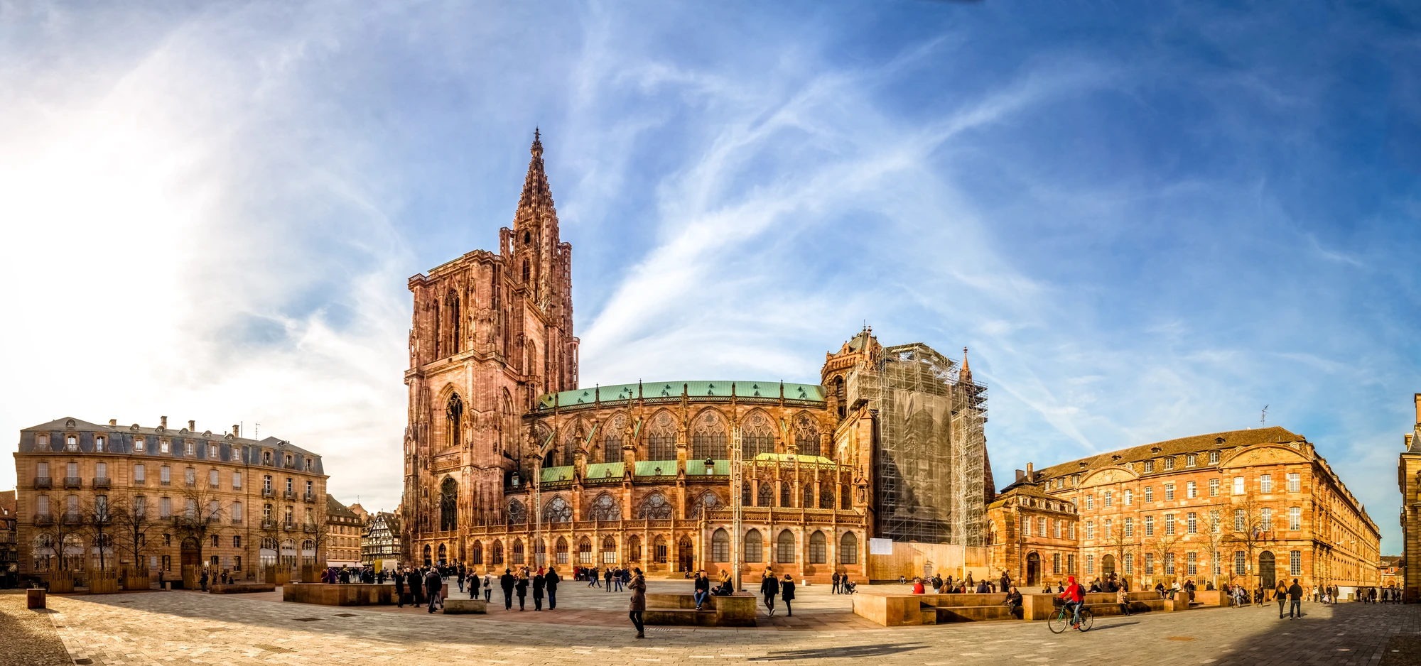 Panoramablick auf das Straßburger Münster, umgeben von der historischen Architektur der Altstadt. Das Bild zeigt lebhafte Aktivitäten auf dem Platz, während die beeindruckende gotische Kathedrale in warmem Licht erstrahlt. Die Bauarbeiten an der rechten Seite sind sichtbar, was die dynamische Atmosphäre des Ortes unterstreicht.