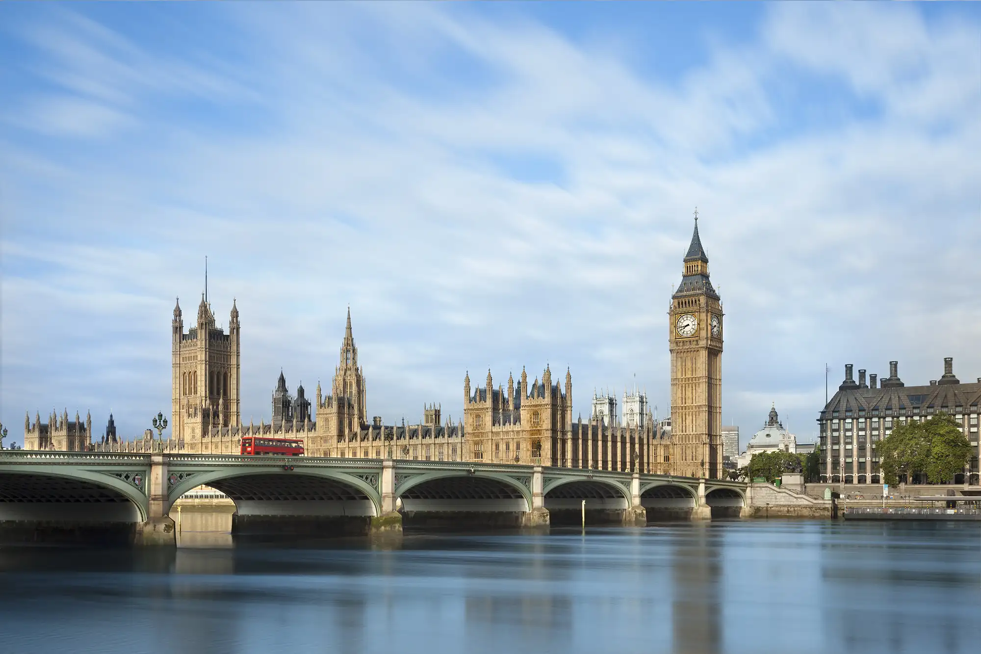 Big Ben, der ikonische Uhrturm, ist ein markantes Wahrzeichen Londons und Teil des Palace of Westminster, wo das britische Parlament sitzt. Die beeindruckende gotische Architektur des Parlamentsgebäudes wird durch die historische Brücke, die über die Themse führt, ergänzt. Diese Ansicht zeigt den ruhigen Fluss und das weiche Licht, das die Szene beleuchtet, während ein typischer roter Doppeldeckerbus über die Brücke fährt, was das urbane Leben Londons widerspiegelt.