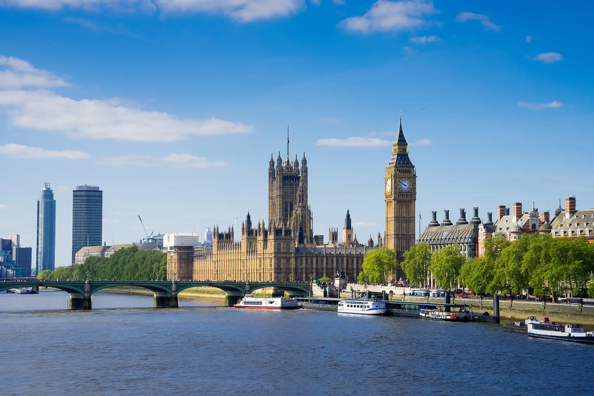 Das Bild zeigt den ikonischen Big Ben und das Houses of Parliament, die an der Themse in London gelegen sind. Die beeindruckende Architektur der beiden Gebäude strahlt historische Bedeutung und zeitlose Eleganz aus. Der Big Ben, der offiziell als Elizabeth Tower bekannt ist, ist mit seiner markanten Uhr und dem hohen Turm ein beliebtes Wahrzeichen der Stadt. Im Vordergrund ist die Westminster Bridge zu sehen, die den Verkehr und Fußgänger über die Themse leitet. Auf der Brücke und entlang des Flusses sind viele Menschen zu erkennen, die die wunderschöne Aussicht genießen. Die grünen Bäume im Vordergrund und der klare blaue Himmel verleihen der Szene eine freundliche und einladende Atmosphäre.