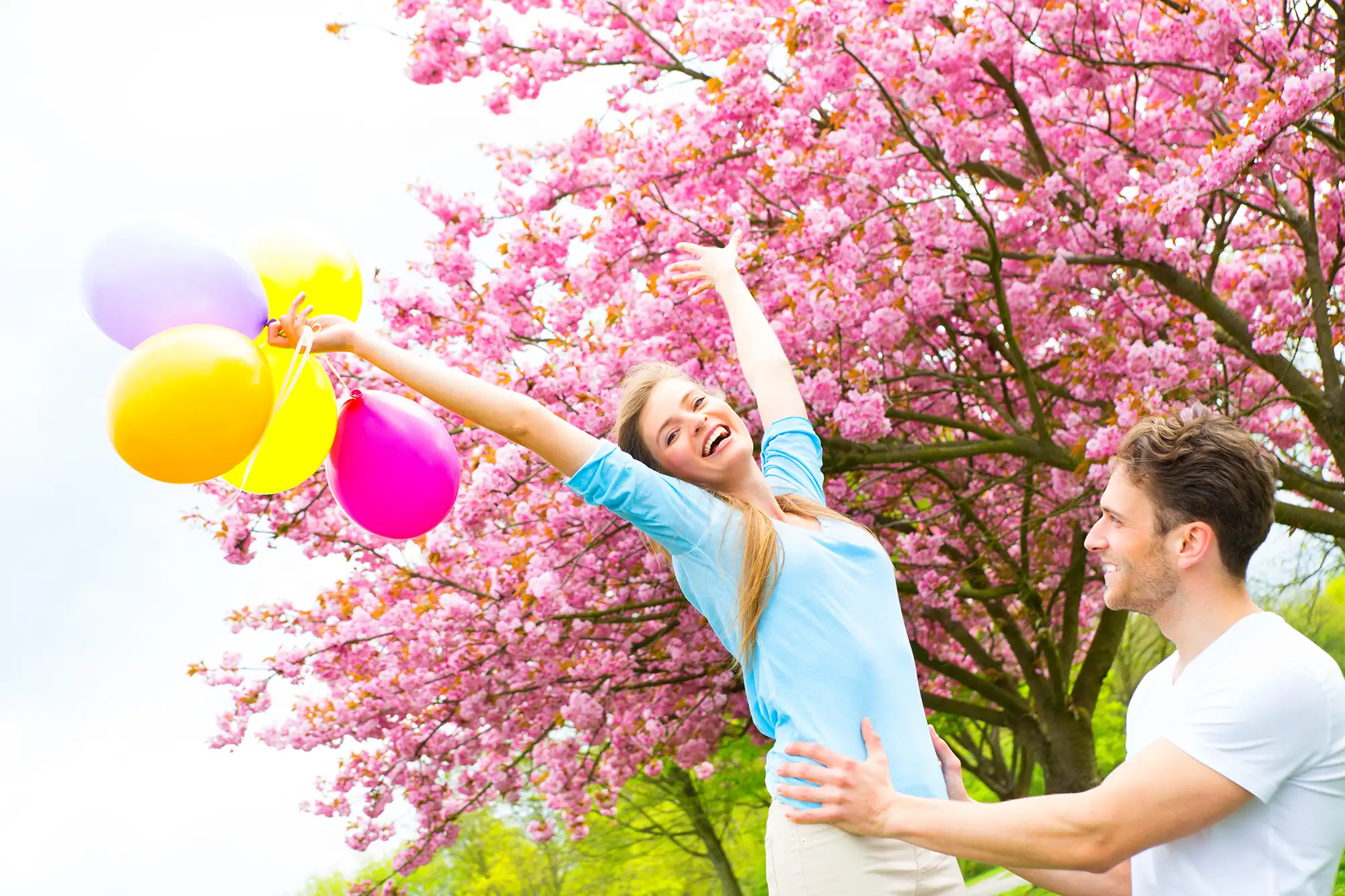 Reisegutscheine verschenken - Mann hebt Frau mit bunten Ballons hoch vor einem Baum mit pinken Blätterm