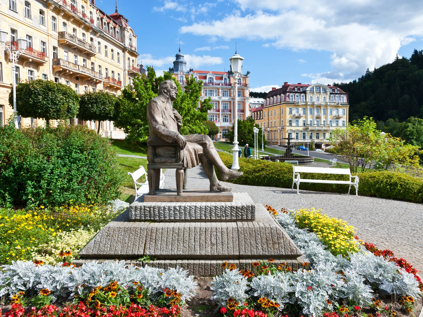 Das Bild zeigt die malerische Kulisse von Marienbad mit einer Statue, die Johann Wolfgang von Goethe gewidmet ist, im Vordergrund. Die Bronzeskulptur, die den Dichter auf einer Bank sitzend darstellt, lädt zum Verweilen ein und erinnert an Goethes Verbindung zu diesem berühmten Kurort. Die Statue ist umgeben von üppigen Blumenbeeten, die mit leuchtenden Farben eine lebendige Atmosphäre schaffen. Im Hintergrund erheben sich prachtvolle historische Gebäude mit kunstvollen Fassaden, die den Charme und die Eleganz von Marienbad verkörpern. Die harmonisch angelegten Gärten und gepflegten Gehwege laden zu entspannten Spaziergängen ein. Der klare, blaue Himmel und das satte Grün der Bäume runden die friedliche Szenerie ab und unterstreichen die einzigartige Kombination aus Natur und Architektur, die Marienbad zu einem beliebten Ziel für Erholungssuchende macht.