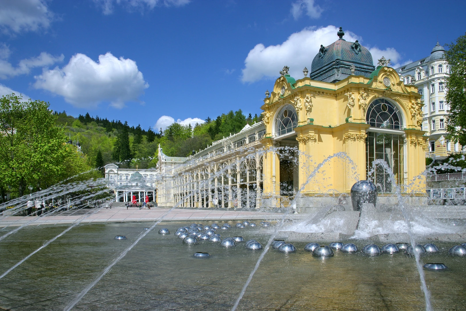 Das Bild zeigt die prachtvolle Hauptkolonnade in Marienbad, ein architektonisches Wahrzeichen des berühmten Kurortes. Die Kolonnade mit ihren filigranen Säulen und kunstvollen Verzierungen erstreckt sich elegant entlang des Platzes. Im Vordergrund beeindruckt die singende Fontäne, die mit ihren Wasserstrahlen ein faszinierendes Schauspiel bietet. Die Strahlen formen kunstvolle Bögen, während glänzende Metallkugeln das Becken schmücken und das Spiel von Licht und Wasser verstärken. Die gelbe Fassade eines angrenzenden Pavillons mit seiner dekorativen Kuppel verleiht der Szene zusätzlichen Glanz und historische Tiefe. Umgeben von saftigem Grün und einer dichten Baumkulisse, spiegelt das Bild die harmonische Verbindung von Natur und Architektur wider. Der strahlend blaue Himmel mit leichten Wolken schafft eine freundliche und einladende Atmosphäre, die den Charme und die Erholung, für die Marienbad bekannt ist, perfekt einfängt.