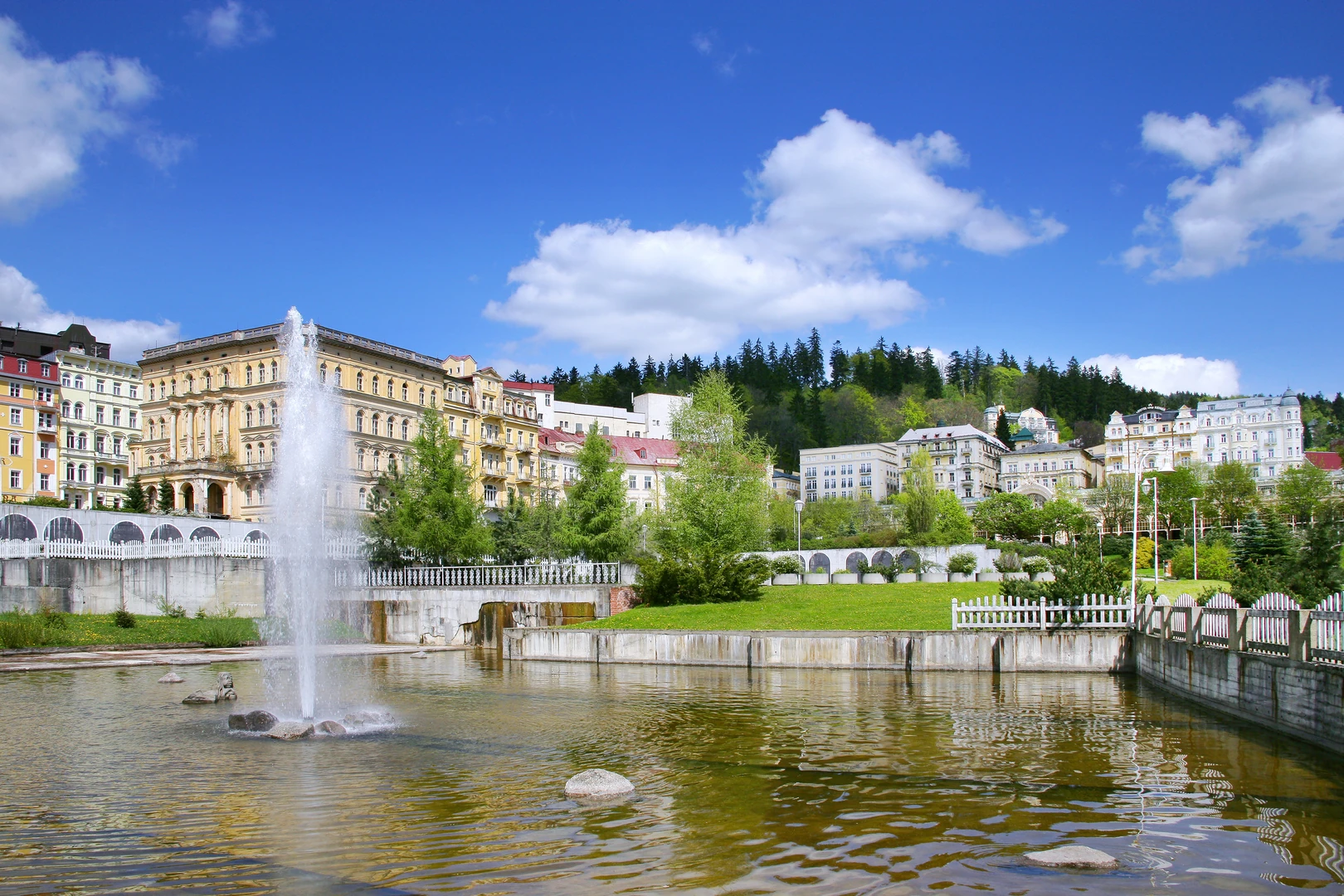 Das Bild zeigt eine idyllische Ansicht von Marienbad, einem der bekanntesten Kurorte Tschechiens. Im Vordergrund sprudelt ein imposanter Springbrunnen, dessen Wasserfontäne kraftvoll in die Höhe schießt. Der Brunnen liegt inmitten eines kleinen Teichs, der von natürlichen Steinen und sauber gestalteten Grünflächen eingerahmt wird. Im Hintergrund erhebt sich eine eindrucksvolle Architektur historischer Gebäude mit aufwendig verzierten Fassaden in sanften Gelb- und Weißtönen. Diese prächtigen Kurhotels und Villen verleihen der Stadt ihren unverkennbaren Charme. Der Horizont ist von üppigen grünen Bäumen geprägt, die die Hügel umgeben und eine malerische Kulisse schaffen. Der klare, blaue Himmel mit leichten Wolken verstärkt die friedliche und erholsame Atmosphäre, die Marienbad als Destination für Wellness und Entspannung so beliebt macht. Dieses Bild vereint den historischen und natürlichen Reichtum der Stadt auf eindrucksvolle Weise.