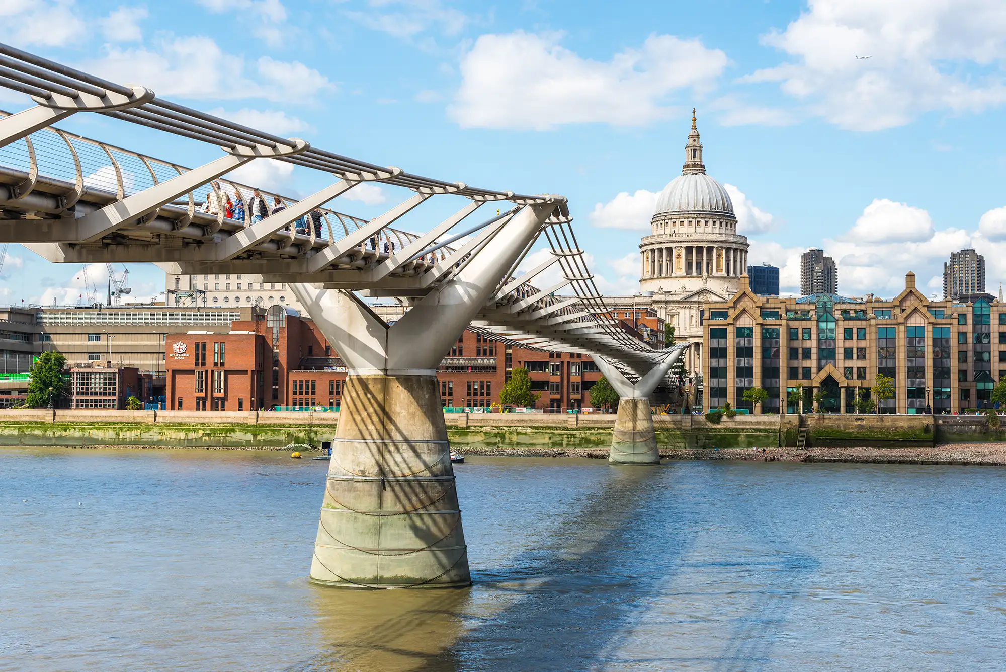 Das Bild zeigt die beeindruckende Millenium Bridge, die sich elegant über die Themse spannt und dabei den Blick auf die markante Kuppel der St. Paul's Cathedral freigibt. Die Brücke, bekannt für ihr modernes Design und ihre Schwingungen, wird von zahlreichen Fußgängern genutzt, die die Verbindung zwischen der City of London und der südlichen Uferseite genießen. Im Hintergrund erhebt sich die St. Paul's Cathedral mit ihrer ikonischen Kuppel, die ein Symbol für Londons reiche Geschichte und Architektur ist. Die Kombination aus der zeitgenössischen Brücke und dem klassischen Gebäude schafft einen faszinierenden Kontrast zwischen alt und neu, was die Vielfalt der Londoner Architektur widerspiegelt. Die klare blaue Himmel und das ruhige Wasser der Themse tragen zur Schönheit der Szene bei, während die lebhaften Farben der Umgebung den pulsierenden Charakter dieser berühmten Stadt unterstreichen.
