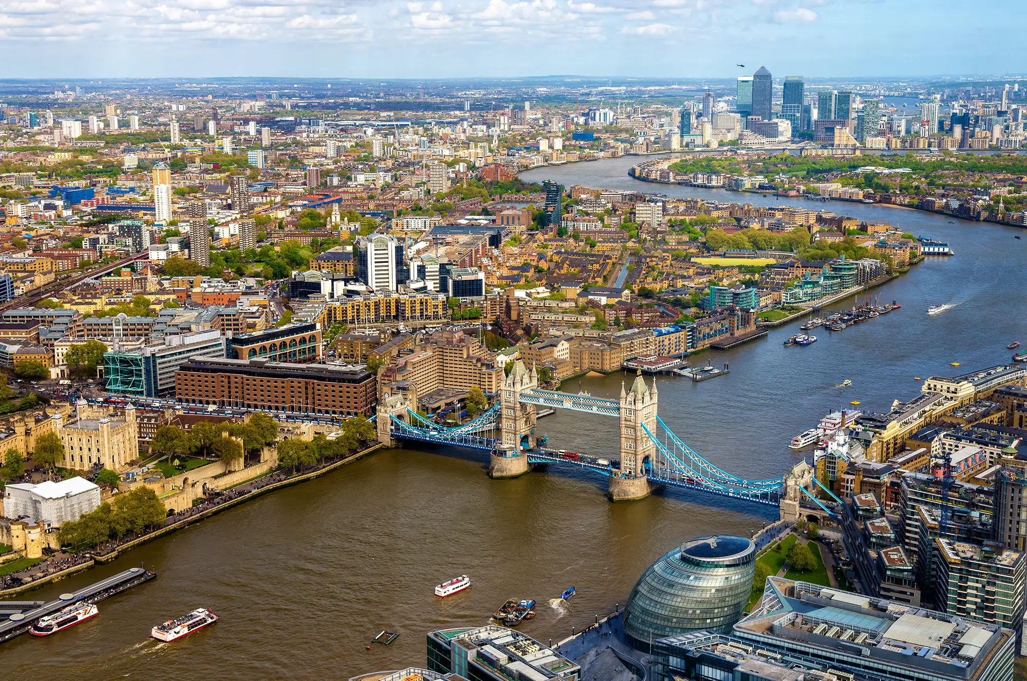 Das Bild zeigt eine beeindruckende Ansicht von London, insbesondere den berühmten Tower Bridge, der majestätisch über die Themse gespannt ist. Die Skyline im Hintergrund ist ein Mix aus modernen Wolkenkratzern und traditionellen Gebäuden, was die Vielfalt und den architektonischen Reichtum der Stadt widerspiegelt. In dieser Perspektive sind auch andere Wahrzeichen wie die Tower of London und verschiedene moderne Bürogebäude sichtbar, die zusammen ein lebendiges Stadtbild schaffen. Die grünen Bereiche und die Wasserwege fügen eine natürliche Komponente hinzu und tragen zur Schönheit dieser urbanen Landschaft bei. Die Gesamtkomposition vermittelt ein Gefühl von Dynamik und Vitalität, typisch für die pulsierende Metropole London.