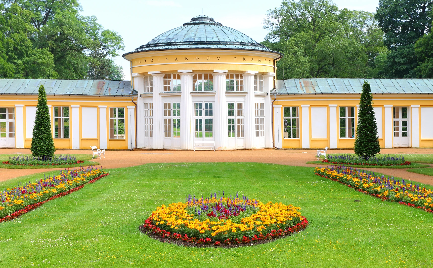 Das Bild zeigt einen malerischen Anblick in Marienbad, Tschechien. Im Zentrum steht ein historischer Pavillon mit der Aufschrift „Ferdinandův pramen“ (Ferdinandsquelle), ein architektonisches Schmuckstück mit hohen weißen Säulen und einer halbrunden Dachkonstruktion. Der Pavillon ist umgeben von gepflegten Grünflächen, die mit kunstvoll arrangierten Blumenbeeten in leuchtenden Farben gestaltet sind. Im Vordergrund sind symmetrisch angeordnete Blumenringe mit gelben, roten und violetten Blüten zu sehen, die dem Ensemble eine harmonische Note verleihen. Die Szenerie wird von einer Kulisse aus hohen, dicht belaubten Bäumen eingerahmt, die Ruhe und Erholung ausstrahlt. Das Bild vermittelt die Eleganz und den historischen Charme von Marienbad als Kurort.