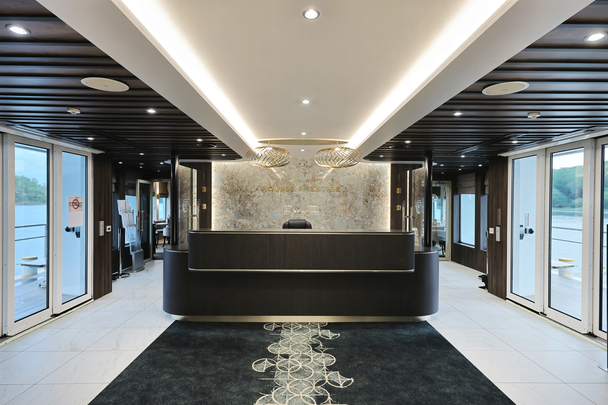 Moderner Empfangstresen mit dunkler Front, darüber zwei runde, goldfarbene Deckenleuchten und eine Wand mit Marmoroptik und goldener Schrift