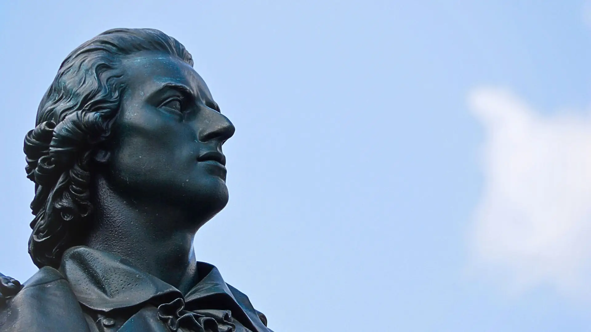 Nahaufnahme der Bronzestatue von Friedrich Schiller mit gewelltem Haar und klassischem Hemd vor blauem Himmel.