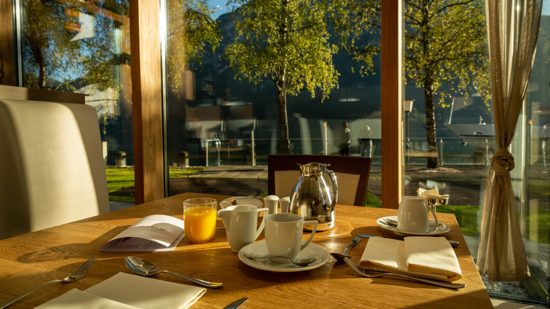 Ein einladender Frühstückstisch im aja Fürstenhaus am Achensee, mit Blick durch große Fenster auf die malerische Natur des Achensees und die umliegenden Bäume, die im goldenen Morgenlicht erstrahlen. Die gemütliche Atmosphäre lädt zum Verweilen ein.