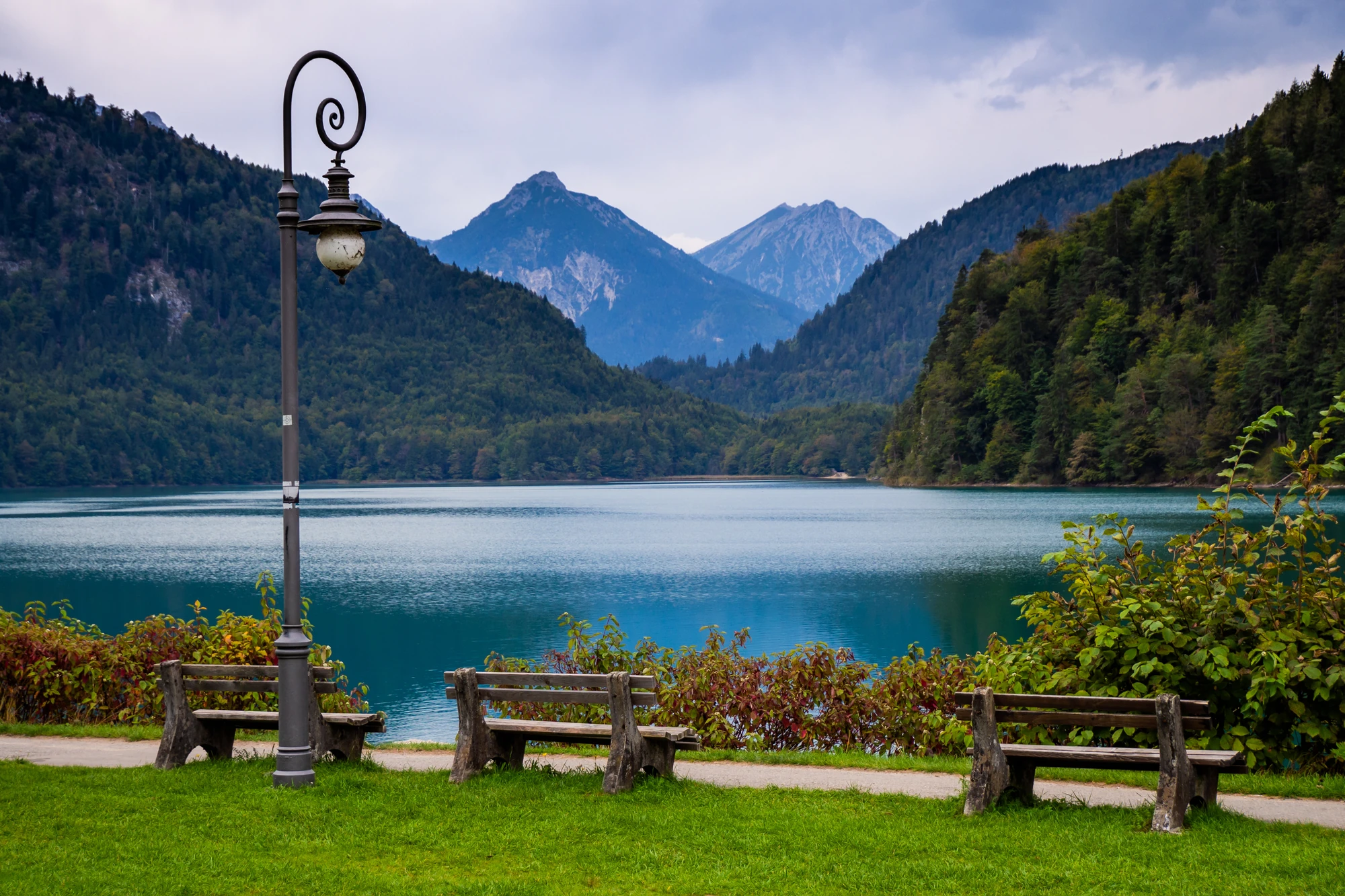 Das Bild zeigt eine idyllische Landschaft in Füssen, mit einem klaren, ruhigen See im Vordergrund, umrahmt von malerischen Bergen. Zwei Holzbanke stehen entlang des Ufers und laden zu einer entspannenden Pause ein, während die sanften Hügel und Wälder im Hintergrund eine friedliche Atmosphäre schaffen. Die Landschaft ist in sanften Herbsttönen gehalten, mit grünen Bäumen und einem dramatisch bewölkten Himmel, der die majestätische Bergkulisse betont.